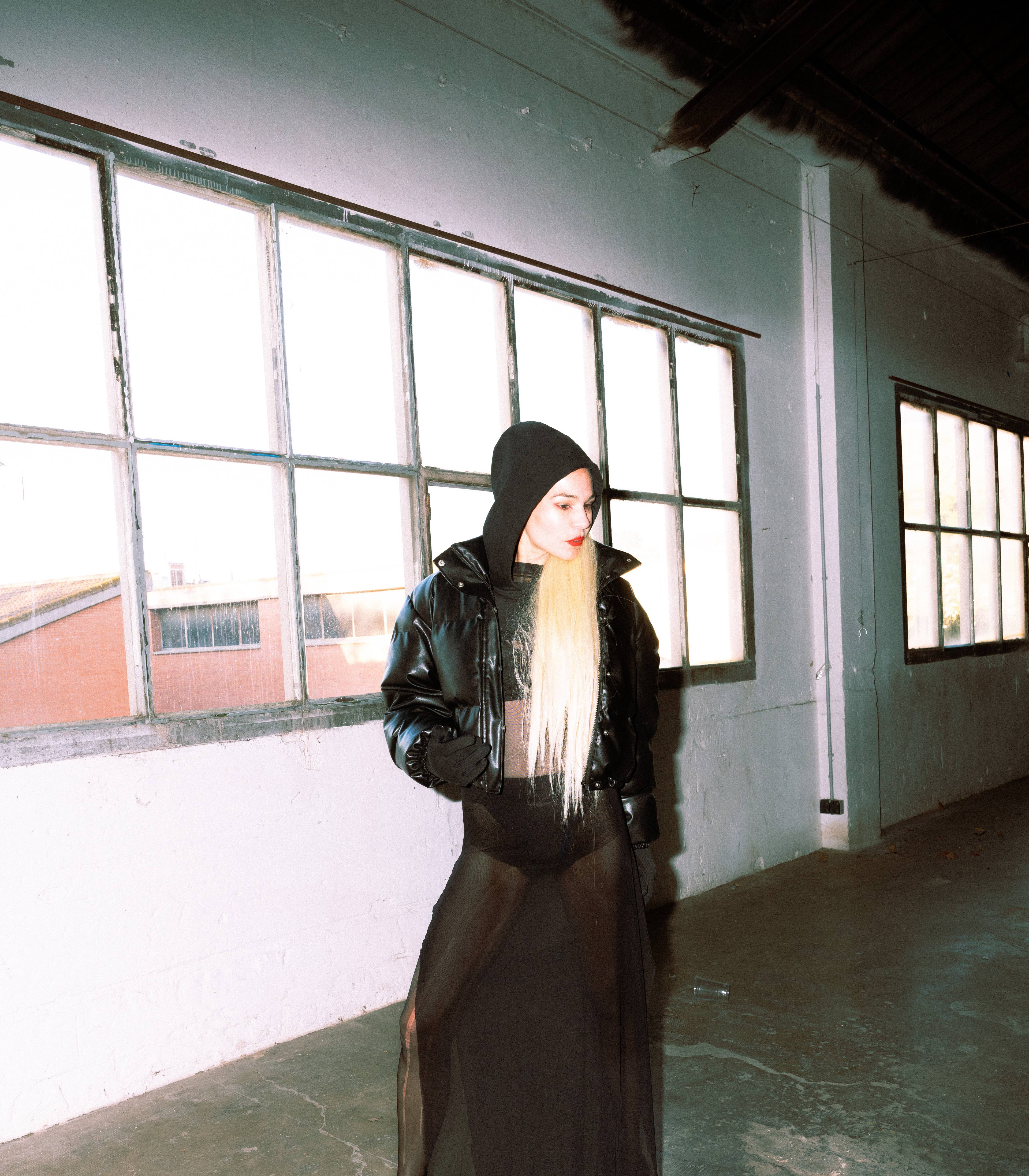 A person with long blond hair and a black hoodie stands in an industrial interior in front of large windows, wearing a sheer black gown and a shiny jacket.