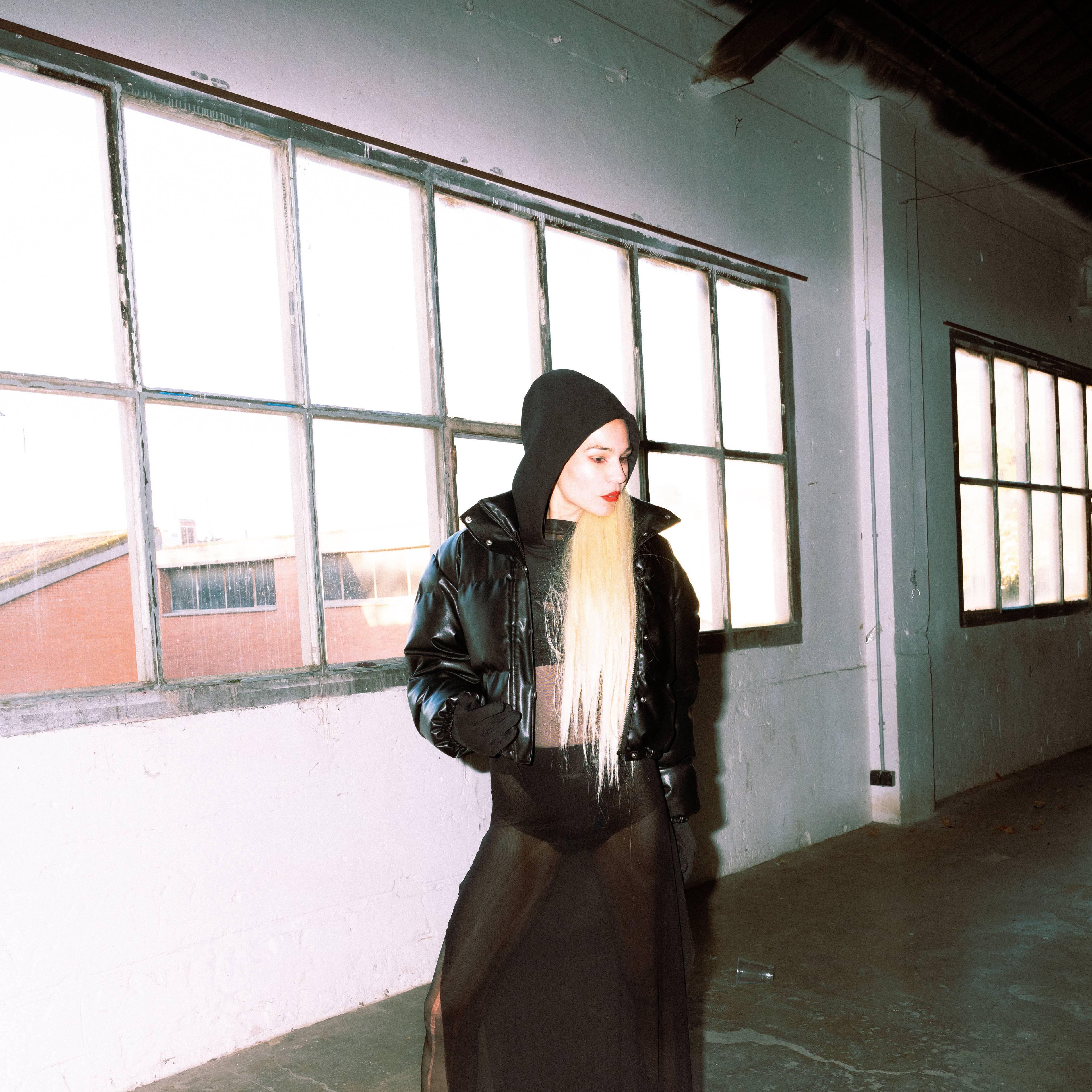 Eine Person mit langen, blonden Haaren und schwarzem Hoodie steht in einem industriellen Innenraum vor großen Fenstern, gekleidet in eine transparente schwarze Robe und eine glänzende Jacke.