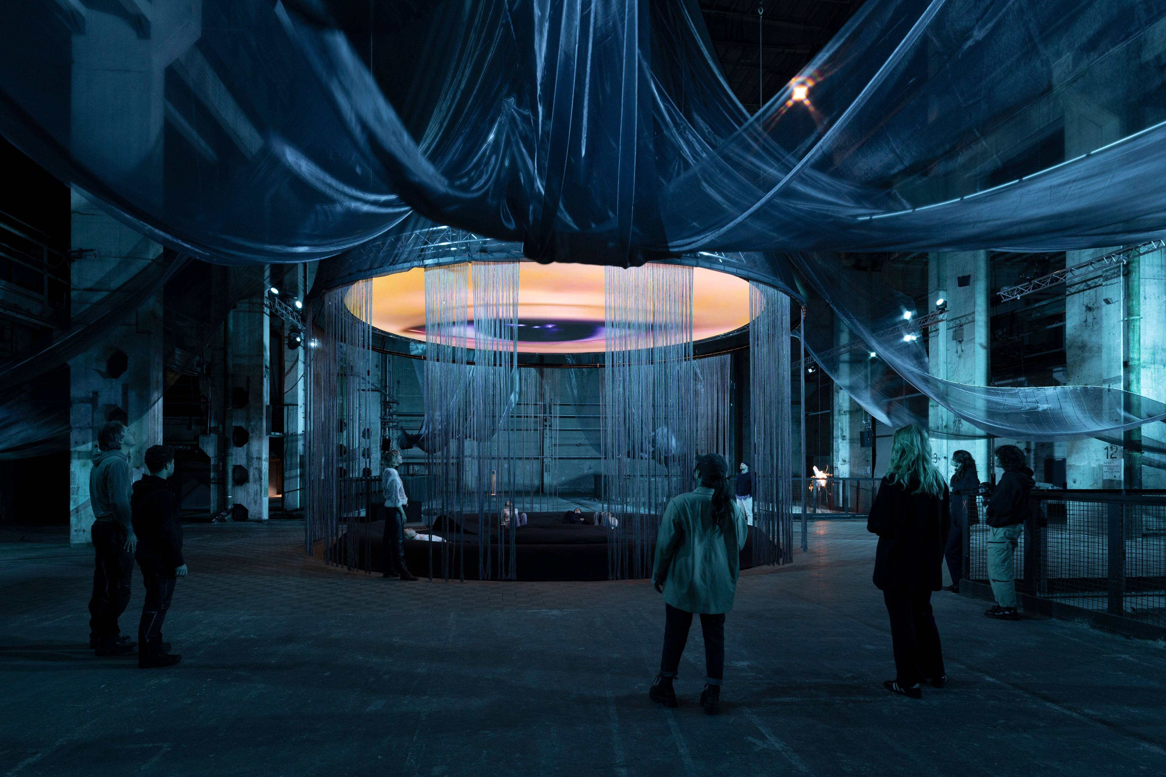 An immersive art installation in an industrial space with a dim, atmospheric blue lighting. A circular structure with hanging threads encloses a central area where people are lying down, gazing at a large illuminated screen above displaying abstract, cosmic imagery in warm hues. Flowing metallic fabric drapes from the ceiling, adding to the ethereal and surreal atmosphere. Several visitors stand around, observing the installation and its elements.