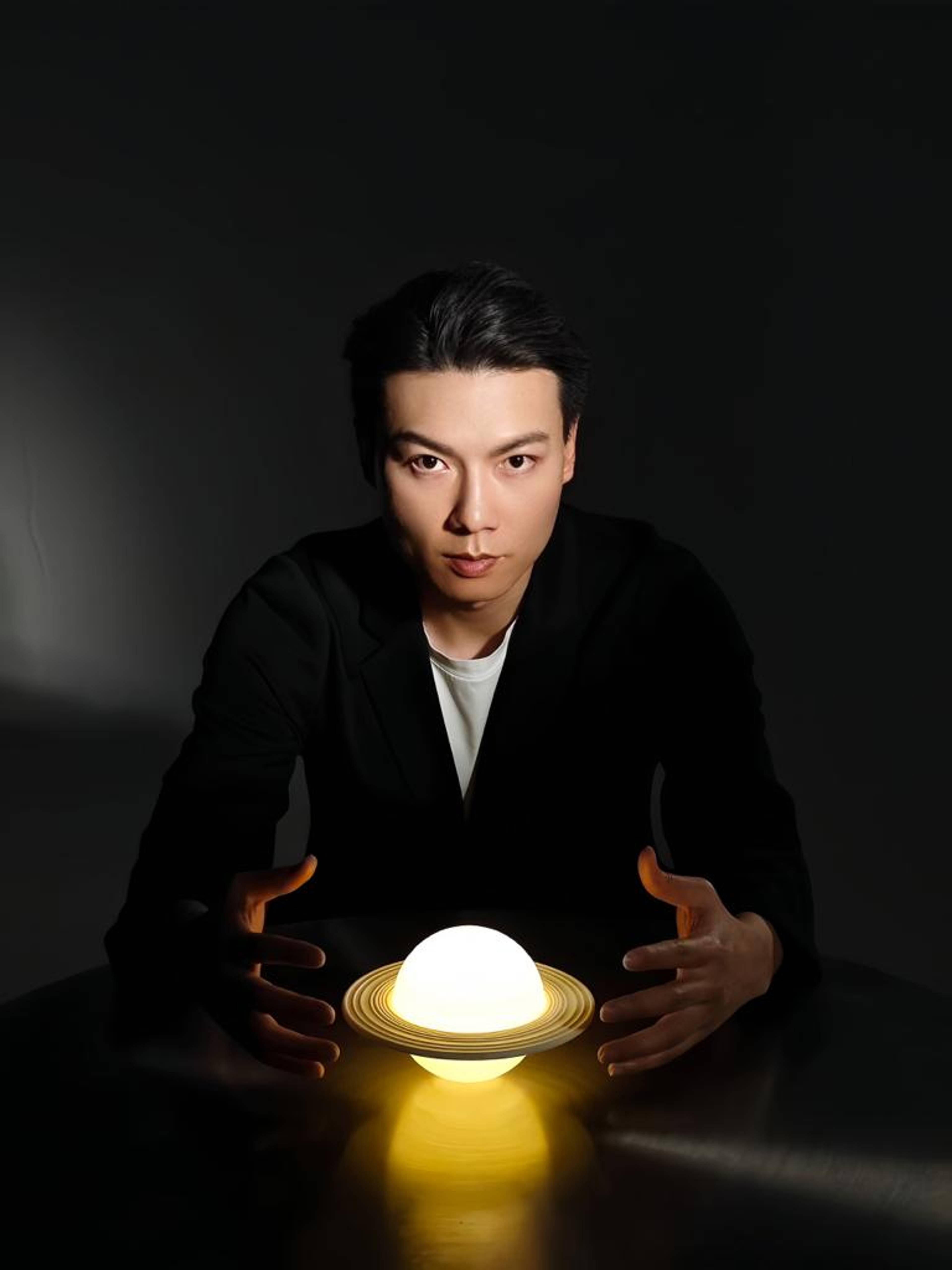 Person in a dark suit seated at a table in a dimly lit room, holding their hands around a glowing spherical lamp with rings resembling the planet Saturn.