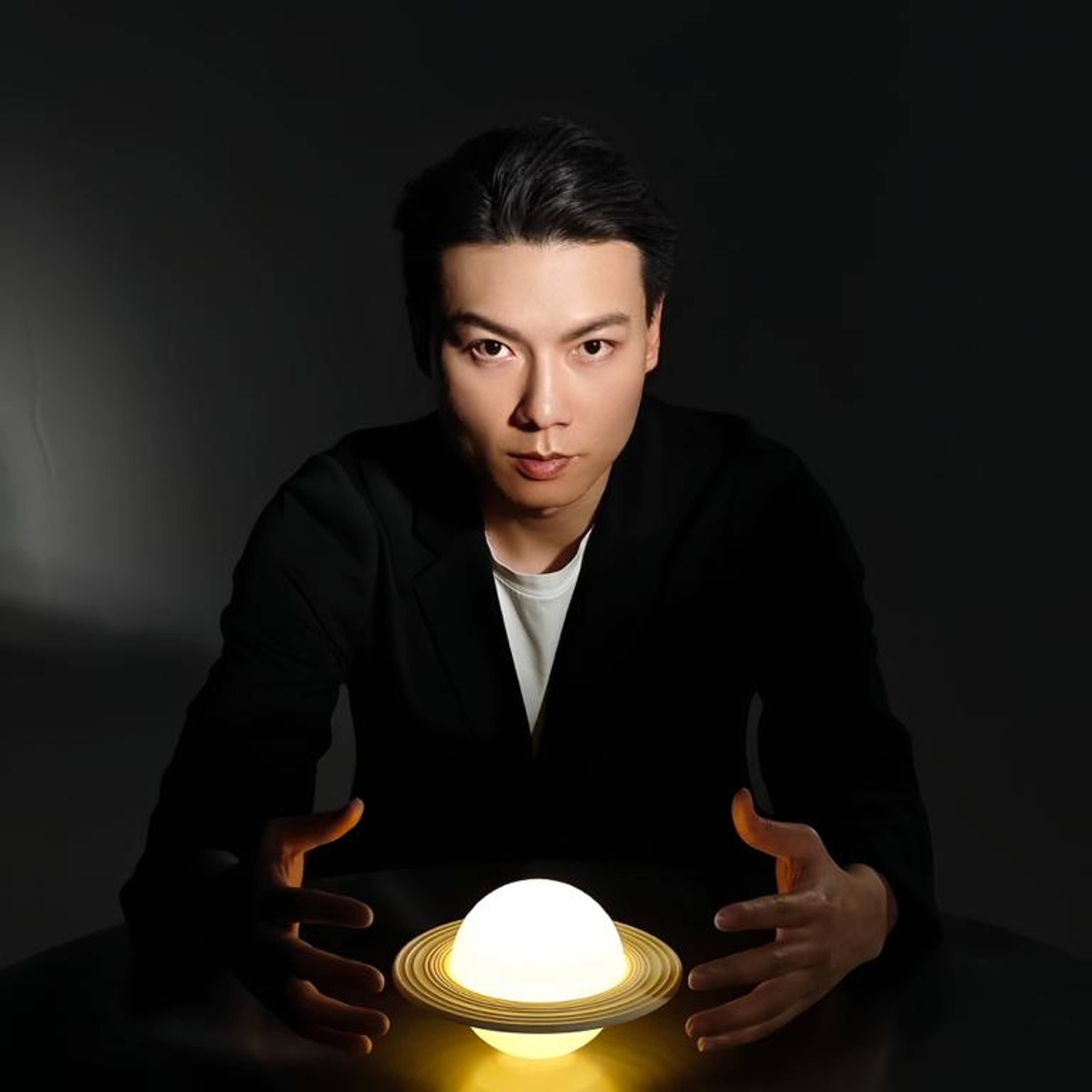 Person in einem dunklen Anzug sitzt an einem Tisch in einem schwach beleuchteten Raum und hält die Hände um eine leuchtende, kugelförmige Lampe mit Ringen, die dem Planeten Saturn ähneln.