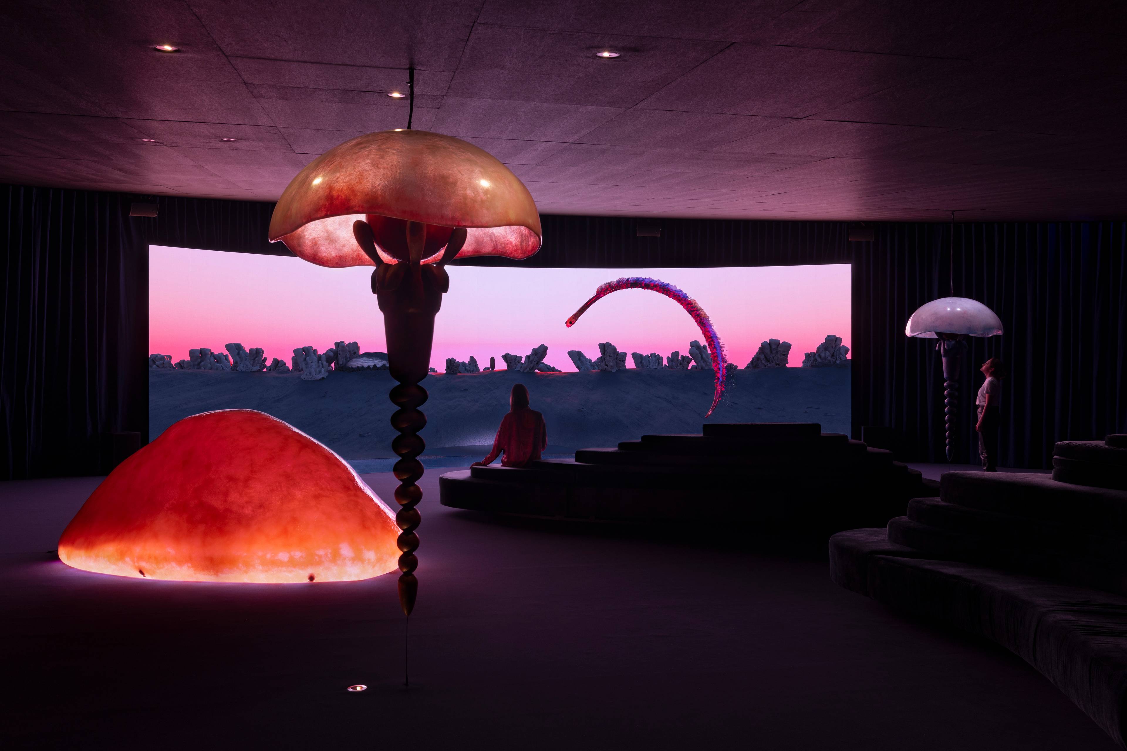 A person sits on tiered seats in a dark room facing a large, curved screen displaying an abstract, futuristic landscape with rocky formations and a pink sky. A large, translucent sculpture hangs from the ceiling in the foreground.