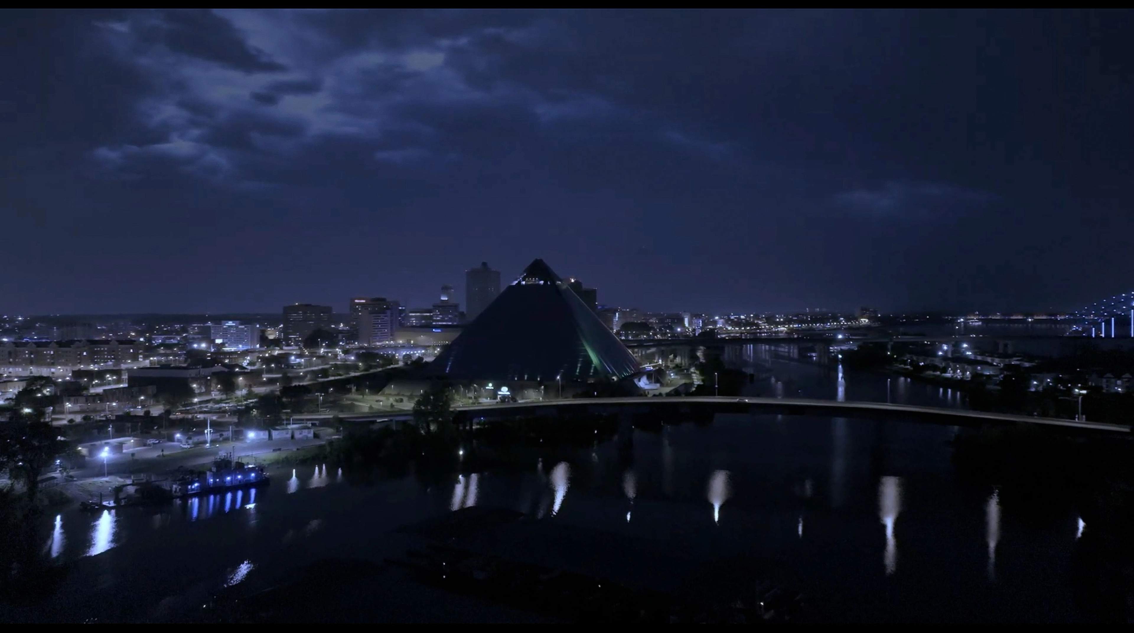 Nachtansicht einer Stadt mit einem großen gläsernen Pyramidengebäude in der Mitte, umgeben von Wasser und Brücken. Stadtlichter spiegeln sich im Fluss unter einem bewölkten Himmel.
