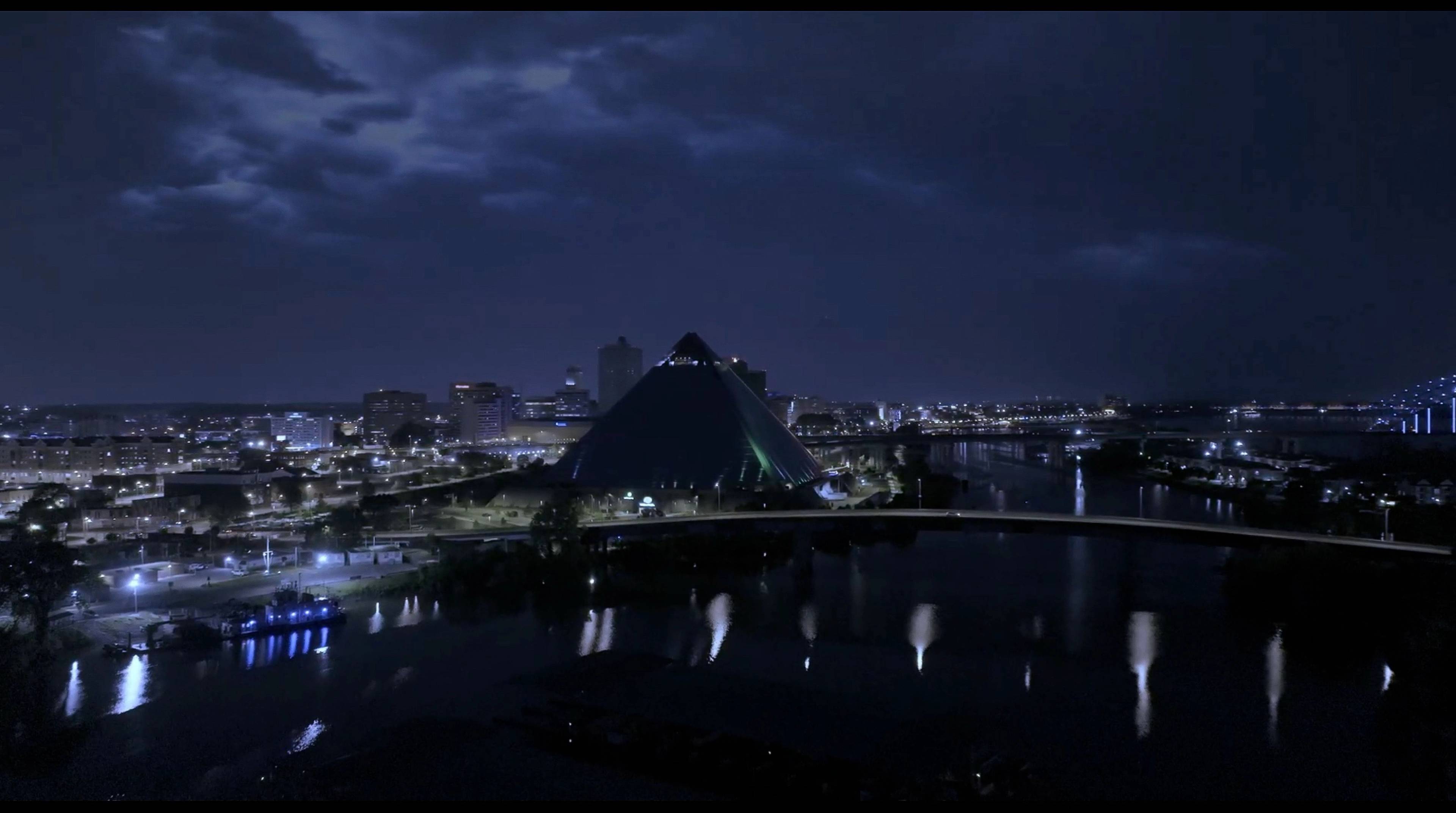 Nachtansicht einer Stadt mit einem großen gläsernen Pyramidengebäude in der Mitte, umgeben von Wasser und Brücken. Stadtlichter spiegeln sich im Fluss unter einem bewölkten Himmel.