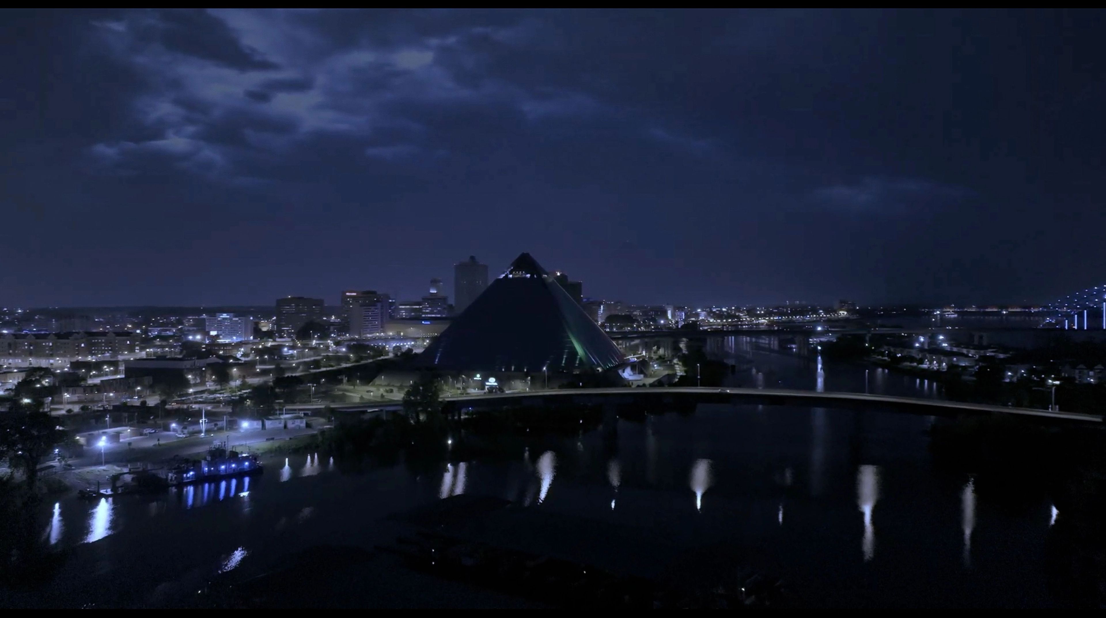 Nachtansicht einer Stadt mit einem großen gläsernen Pyramidengebäude in der Mitte, umgeben von Wasser und Brücken. Stadtlichter spiegeln sich im Fluss unter einem bewölkten Himmel.