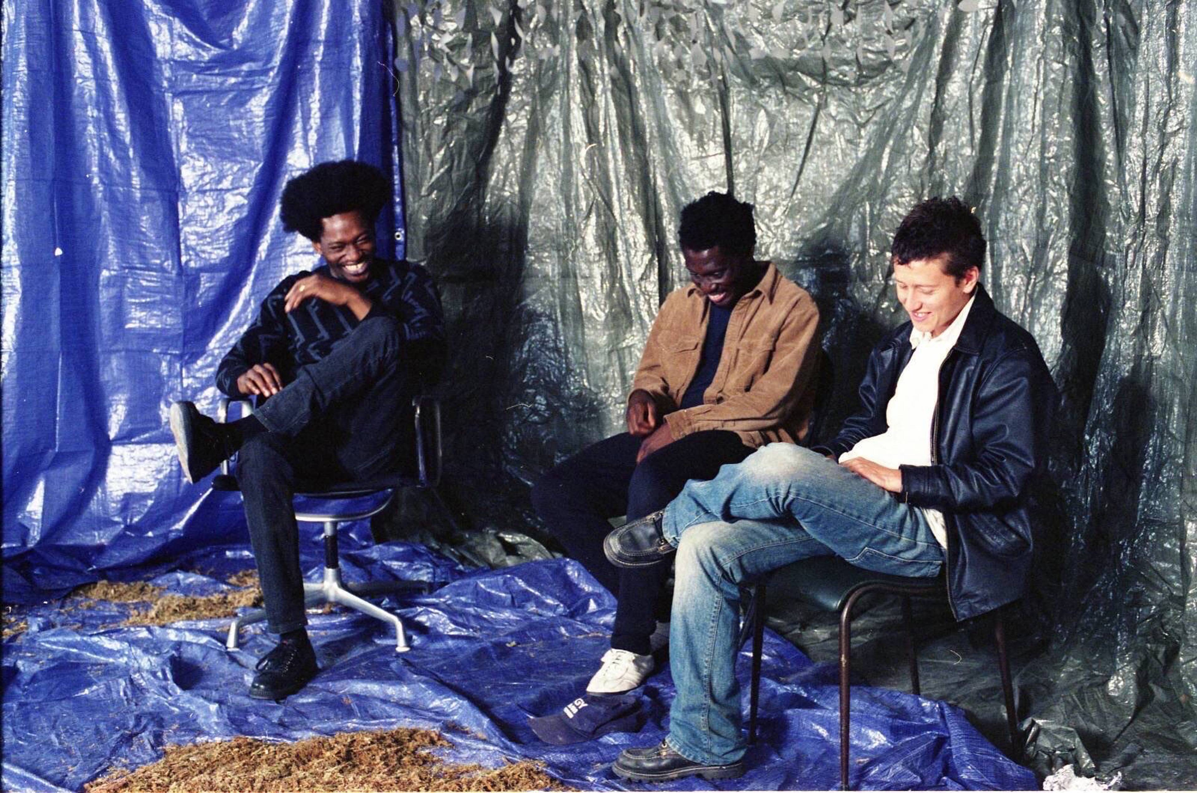 Die CURL-Kollektivmitglieder Mica Levi, Brother May und Coby Sey sitzen lächelnd vor blauen und schwarzen Planen.