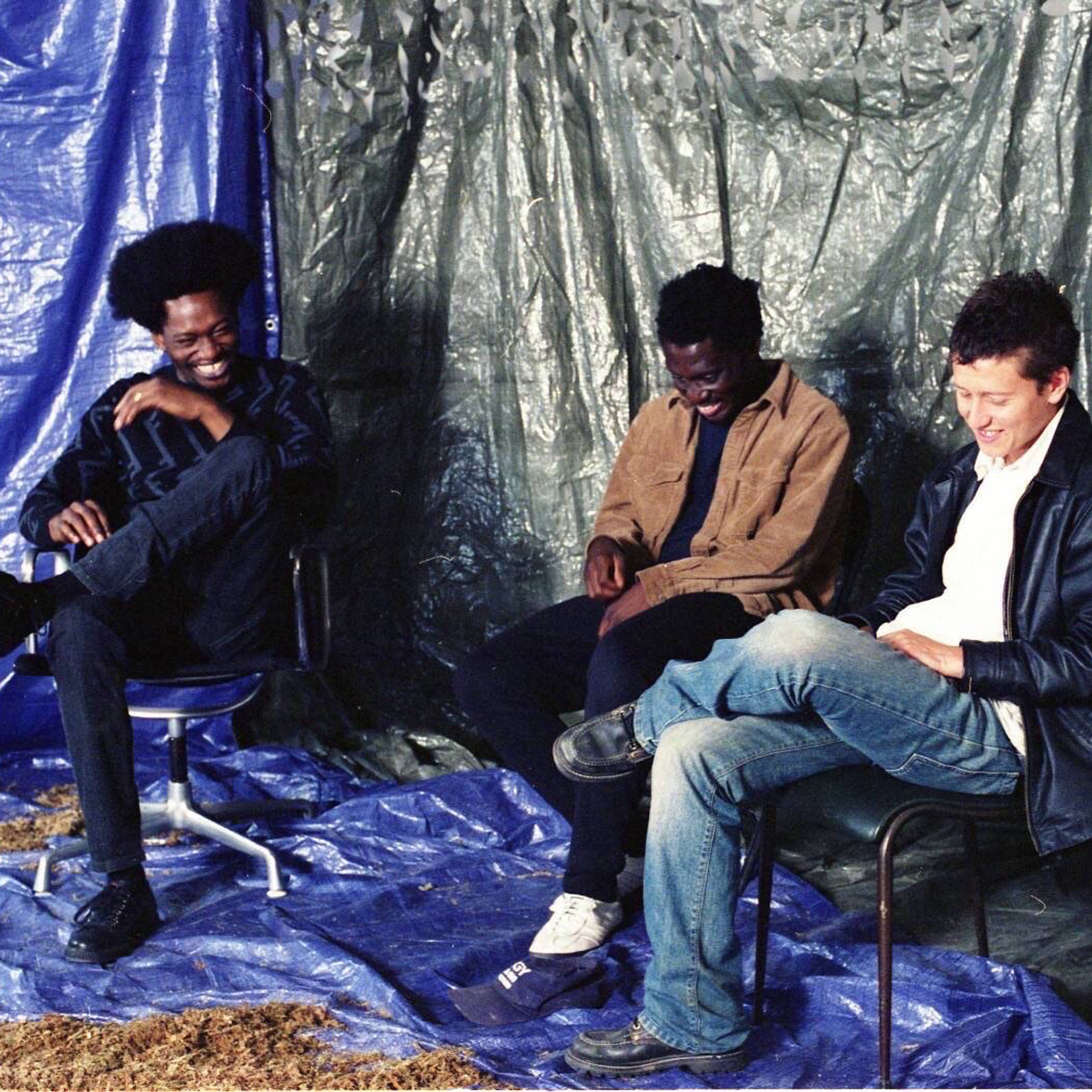 Die CURL-Kollektivmitglieder Mica Levi, Brother May und Coby Sey sitzen lächelnd vor blauen und schwarzen Planen.