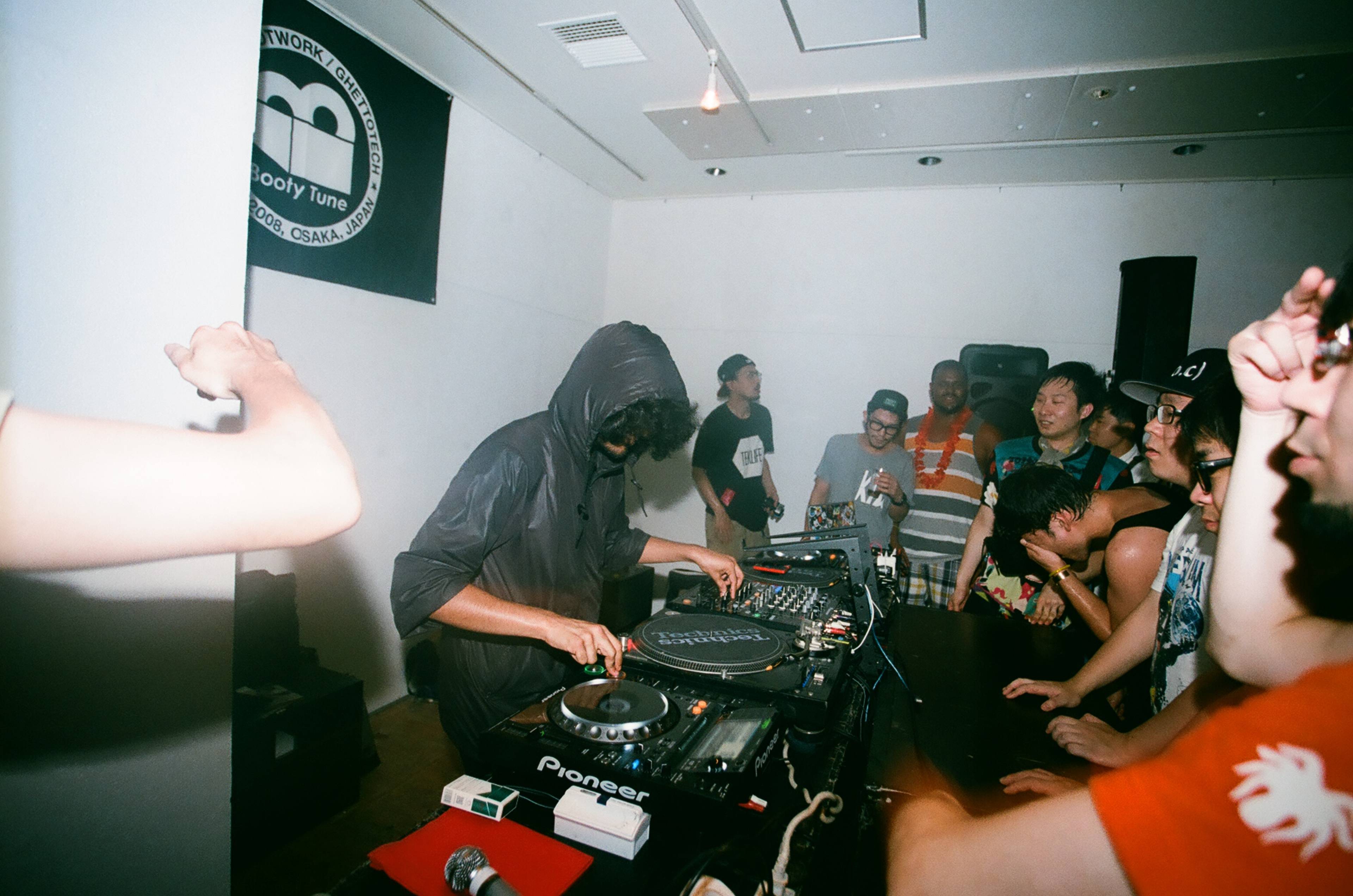 A DJ wearing a hoodie mixes music on turntables in a crowded, lively room. People around the DJ booth are smiling, dancing, and enjoying the music. A banner with a logo is on the wall, and various equipment is visible on the table.