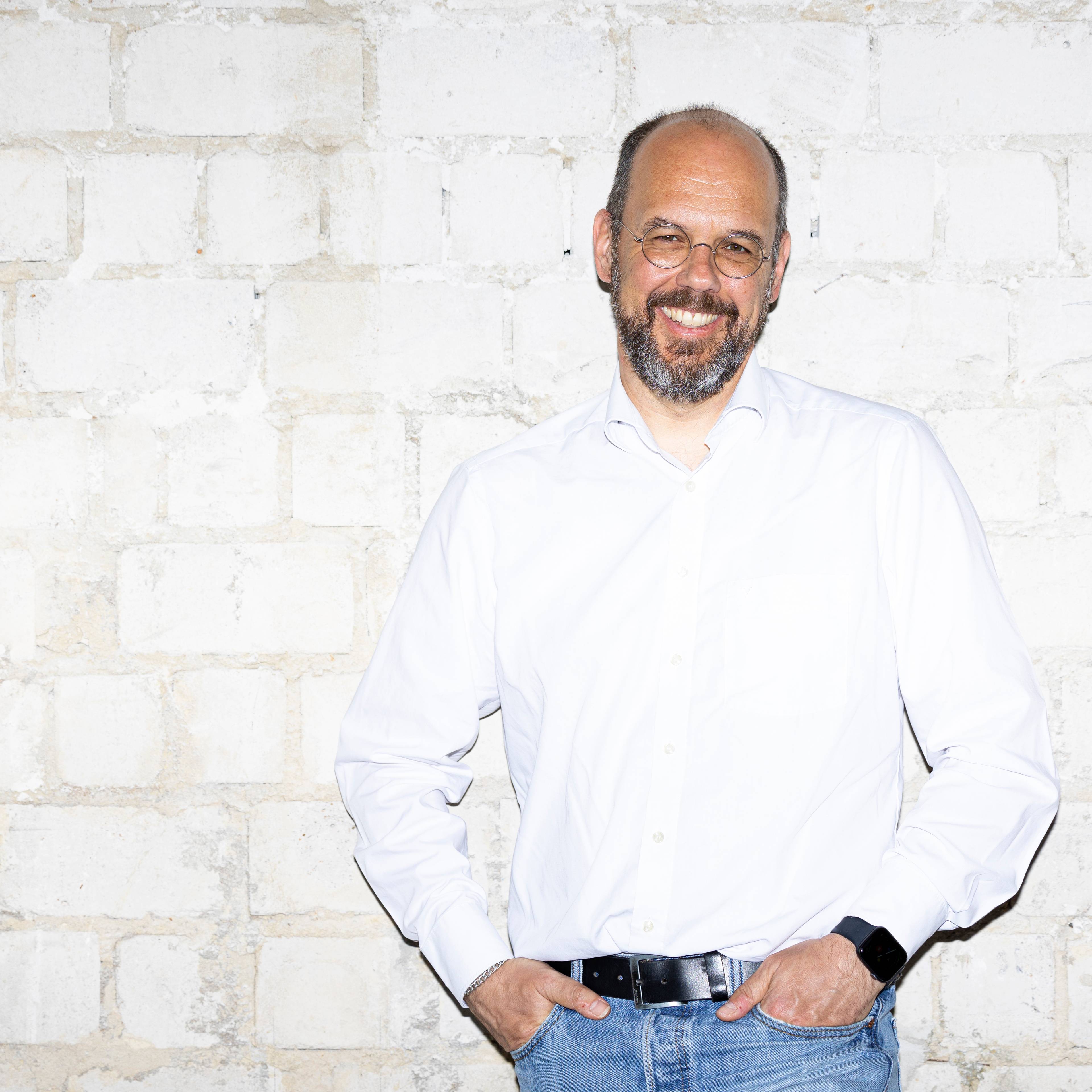 A smiling man with glasses, a beard, and a white shirt stands in front of a white-painted brick wall. He is wearing jeans and has his hands in his pockets.