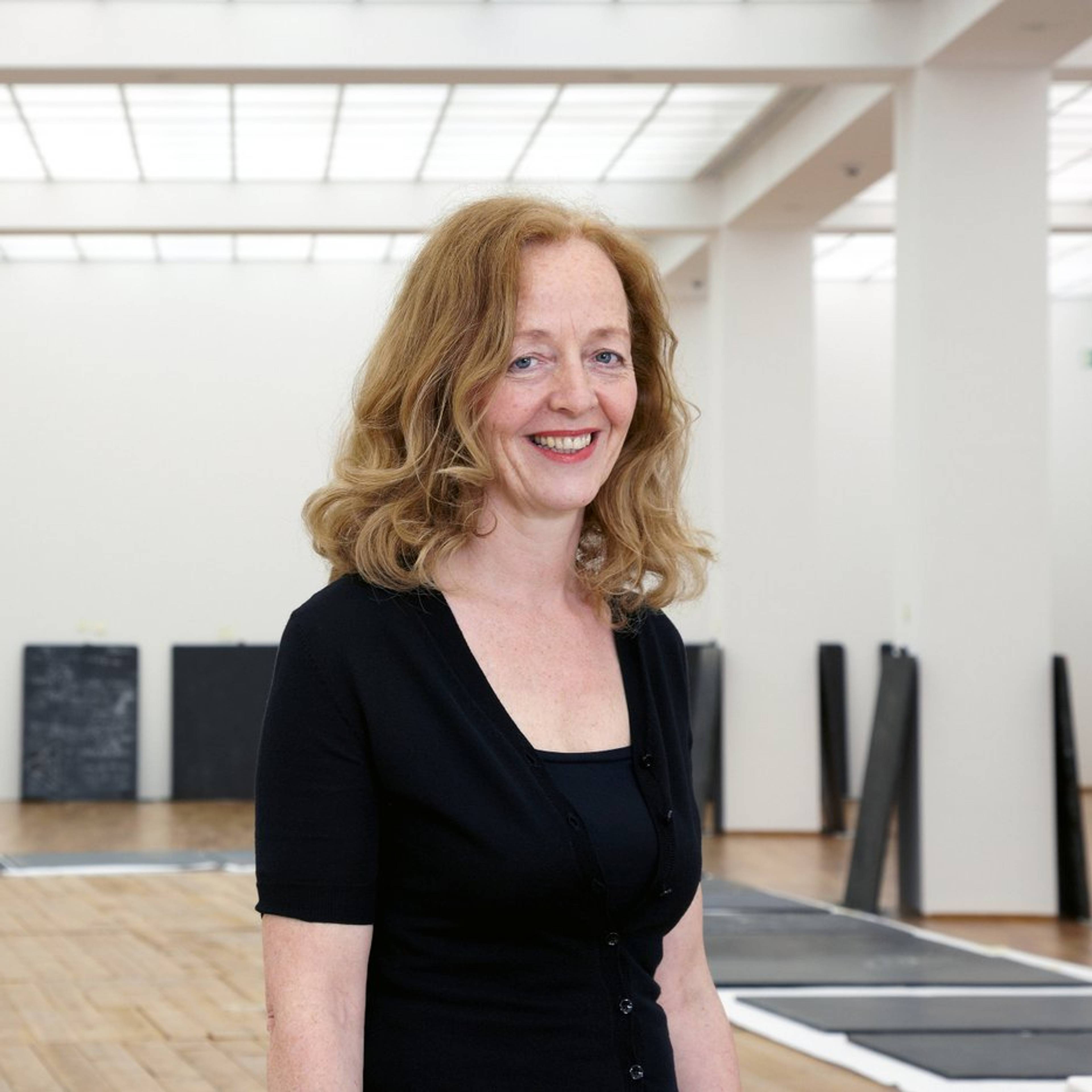 A woman with shoulder-length blonde hair, wearing a black top, smiles in an art gallery with white walls and wooden floors. Black and white artworks are displayed in the background.