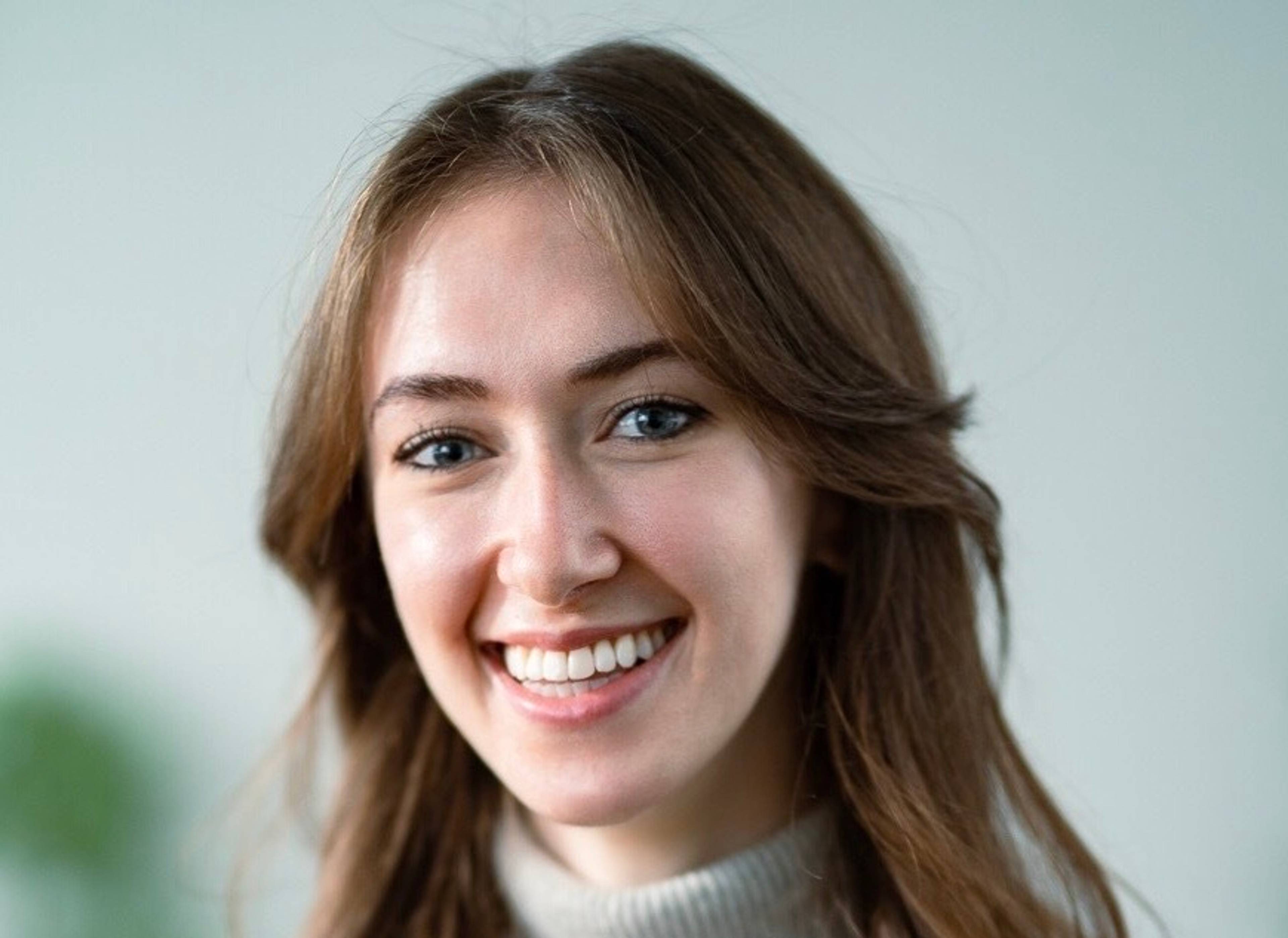 Porträt einer lachenden Person mit langen braunen Haaren und hellem Rollkragenpullover vor einem neutralen Hintergrund.