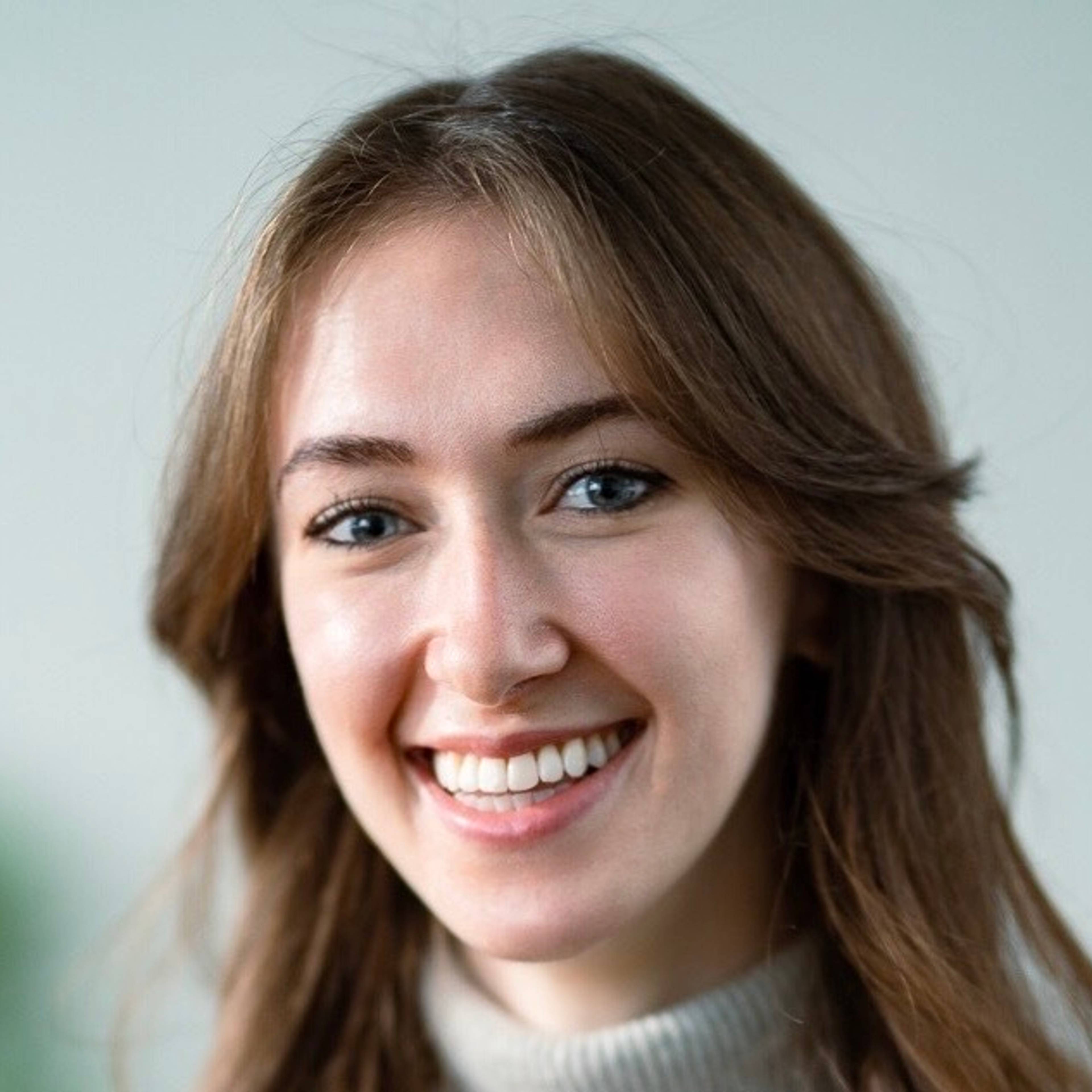 Portrait of a smiling person with long brown hair and a light turtleneck sweater against a neutral background.