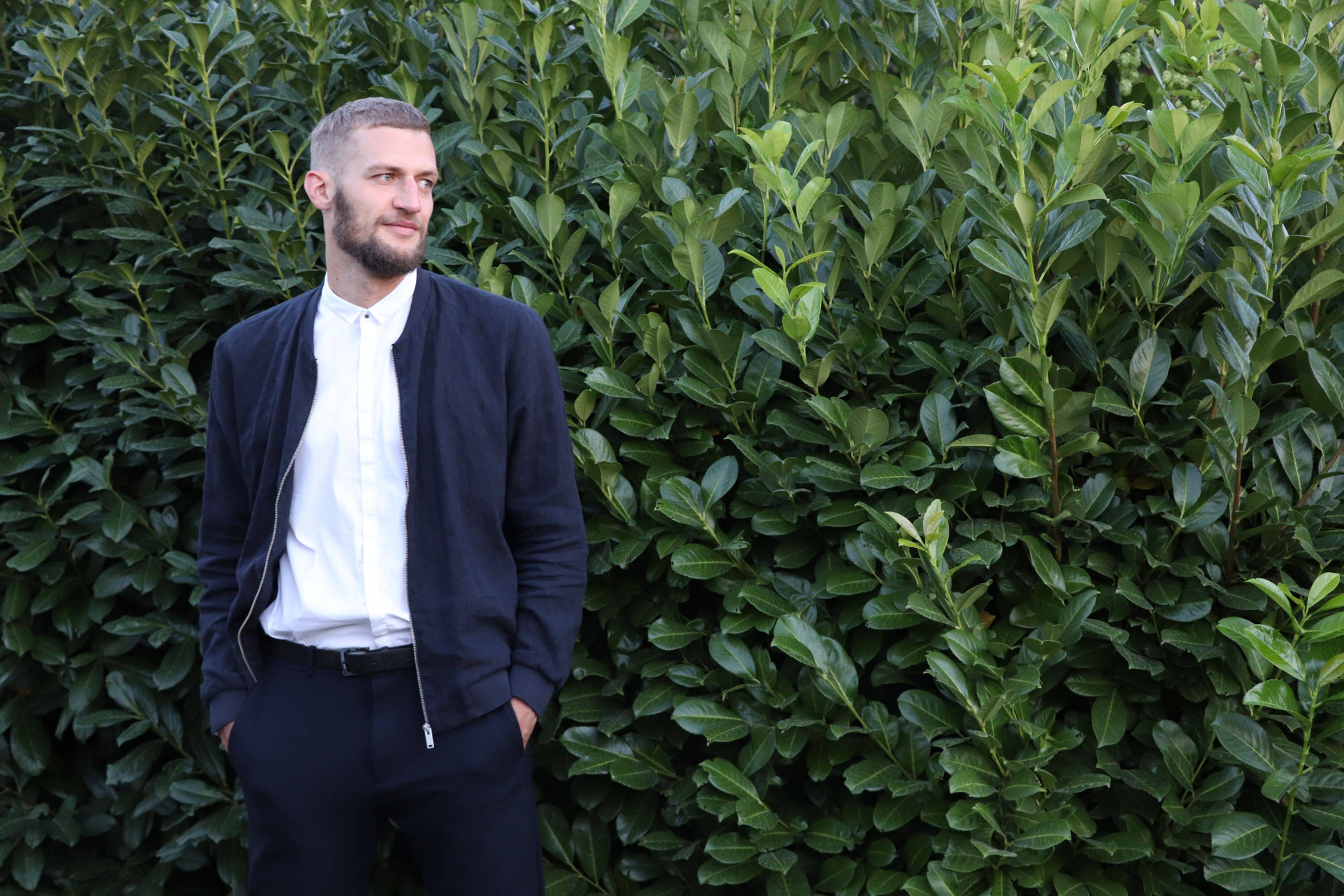 A man with a beard stands in front of dense green foliage, wearing a white shirt, dark jacket, and pants.