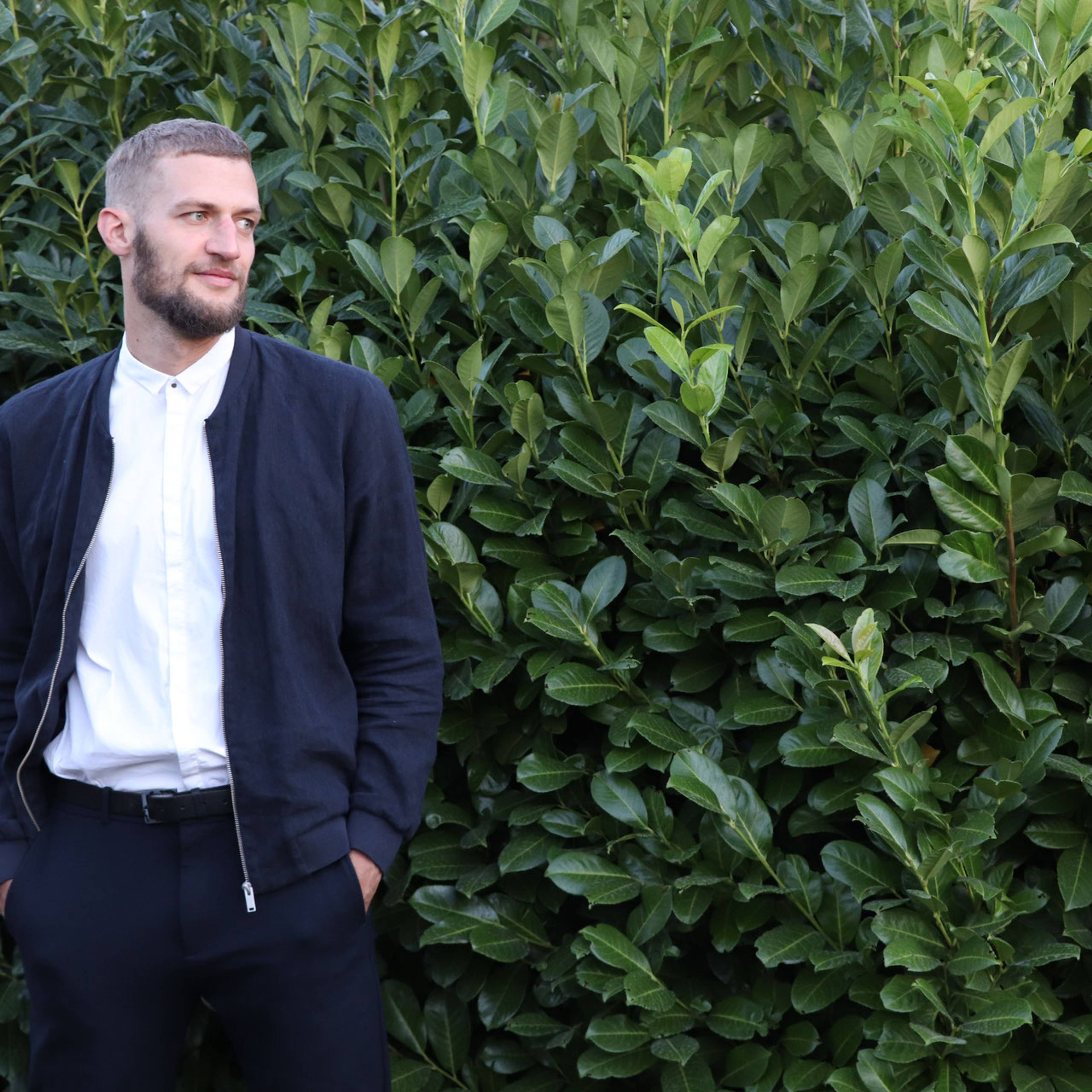 A man with a beard stands in front of dense green foliage, wearing a white shirt, dark jacket, and pants.