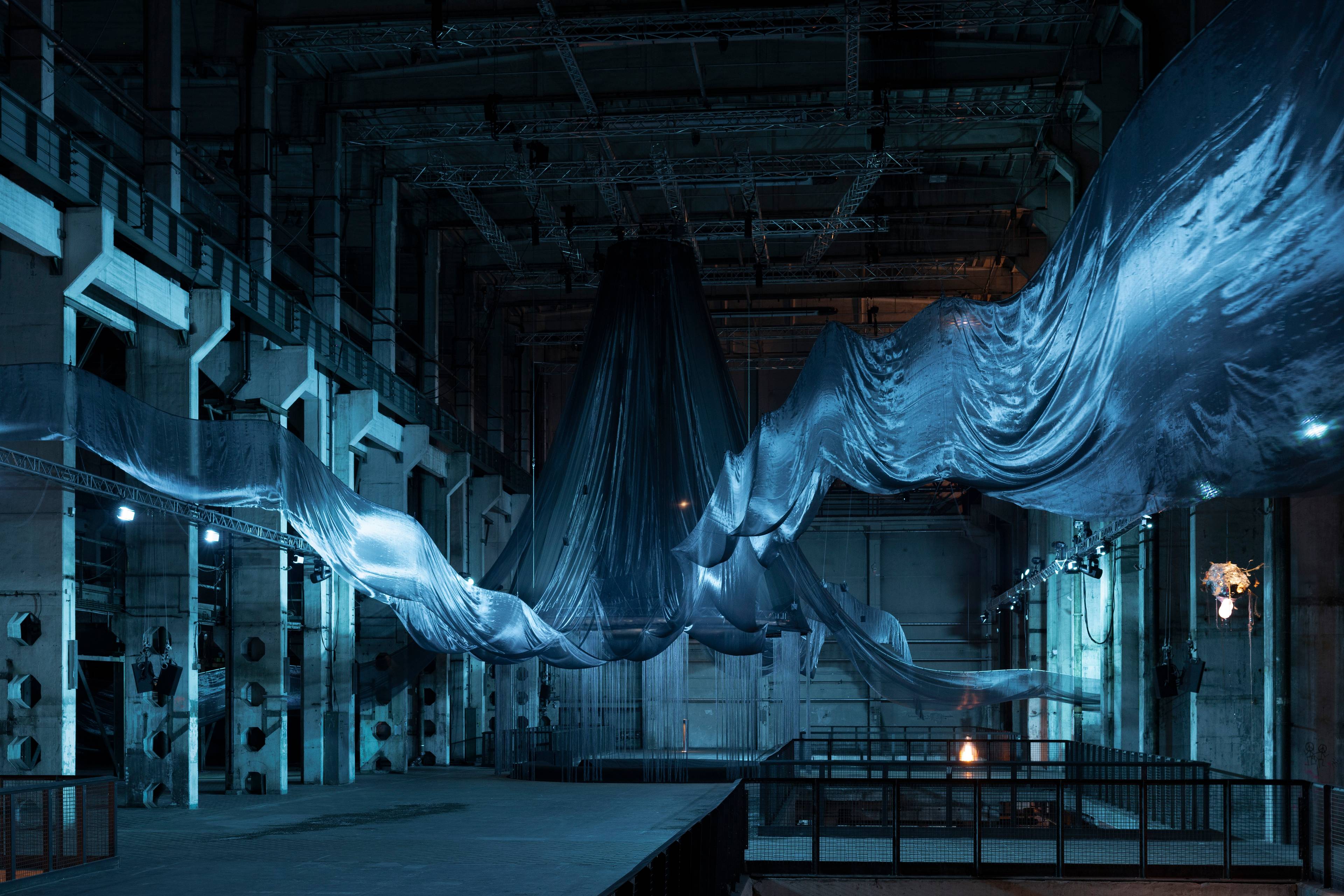 A large-scale art installation in an industrial space features flowing, semi-transparent black fabric suspended from the ceiling. The installation is bathed in a cool blue light, casting eerie reflections on the fabric and surrounding architecture. The dramatic drapery extends across the space, creating a dynamic, immersive atmosphere. A small illuminated structure hangs on the right side, adding a mysterious focal point.