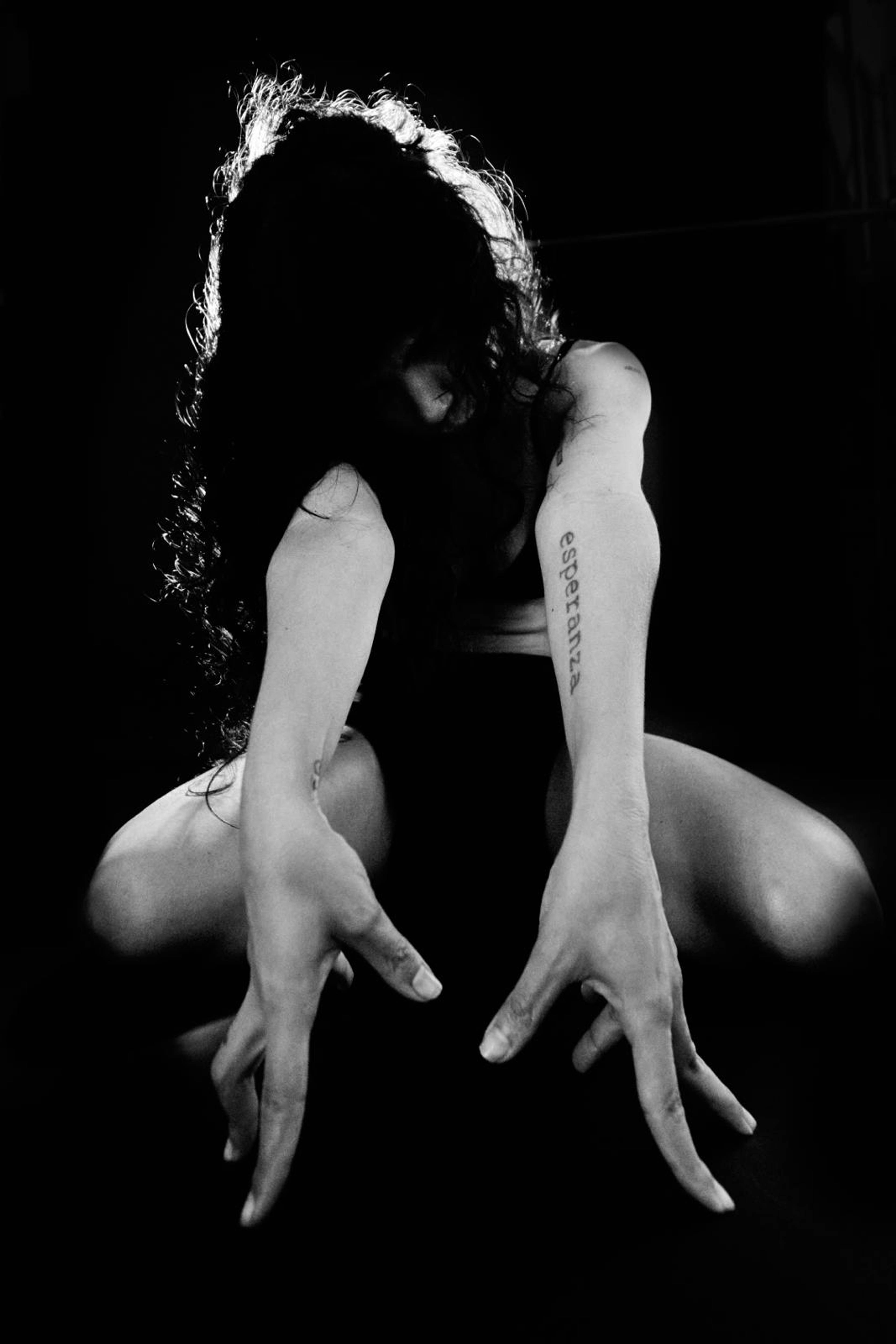 Black and white photo of a person with long hair, crouching with arms extended towards the camera. The word esperanza is tattooed on their arm. The lighting creates dramatic shadows, highlighting the expressive pose.