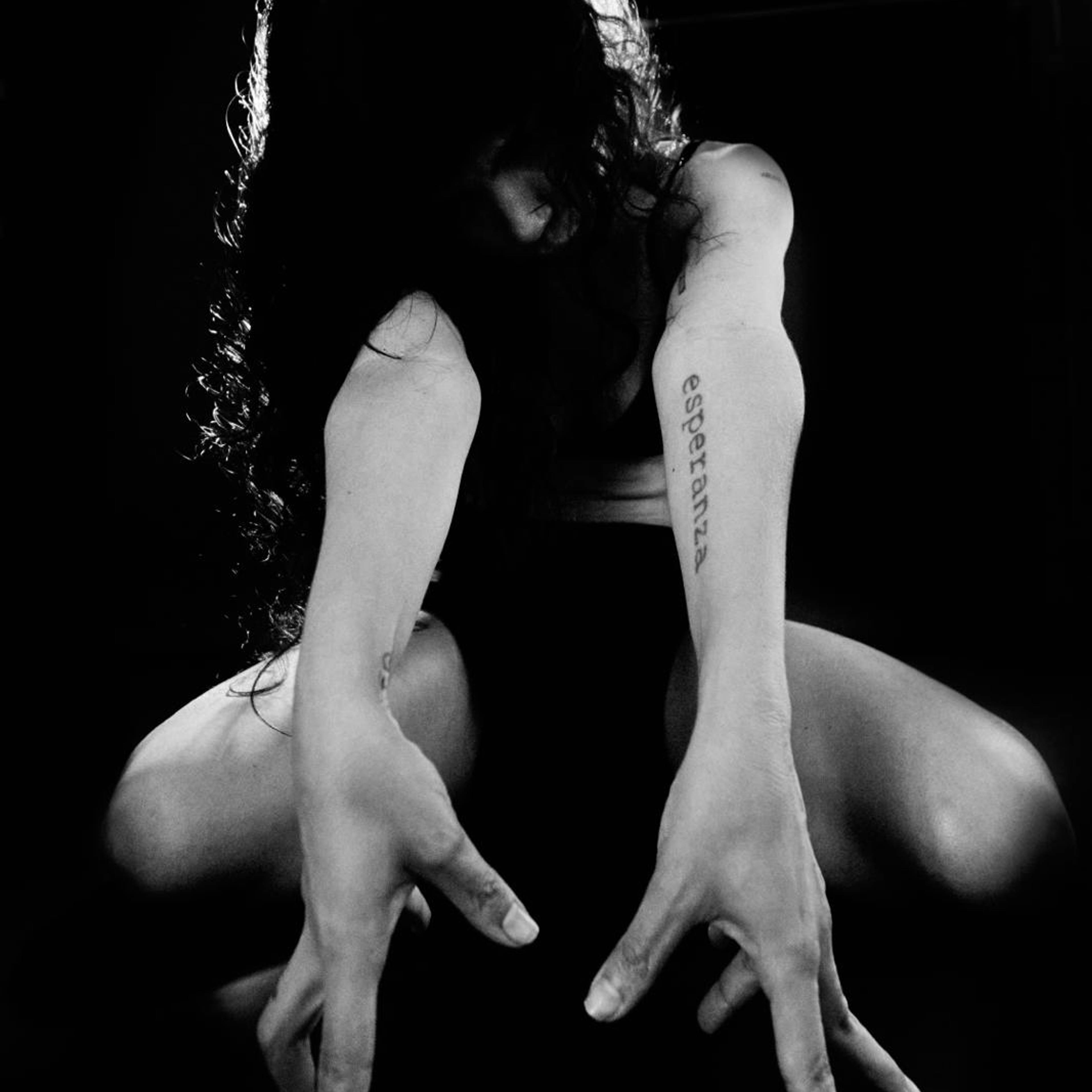 Black and white photo of a person with long hair, crouching with arms extended towards the camera. The word esperanza is tattooed on their arm. The lighting creates dramatic shadows, highlighting the expressive pose.
