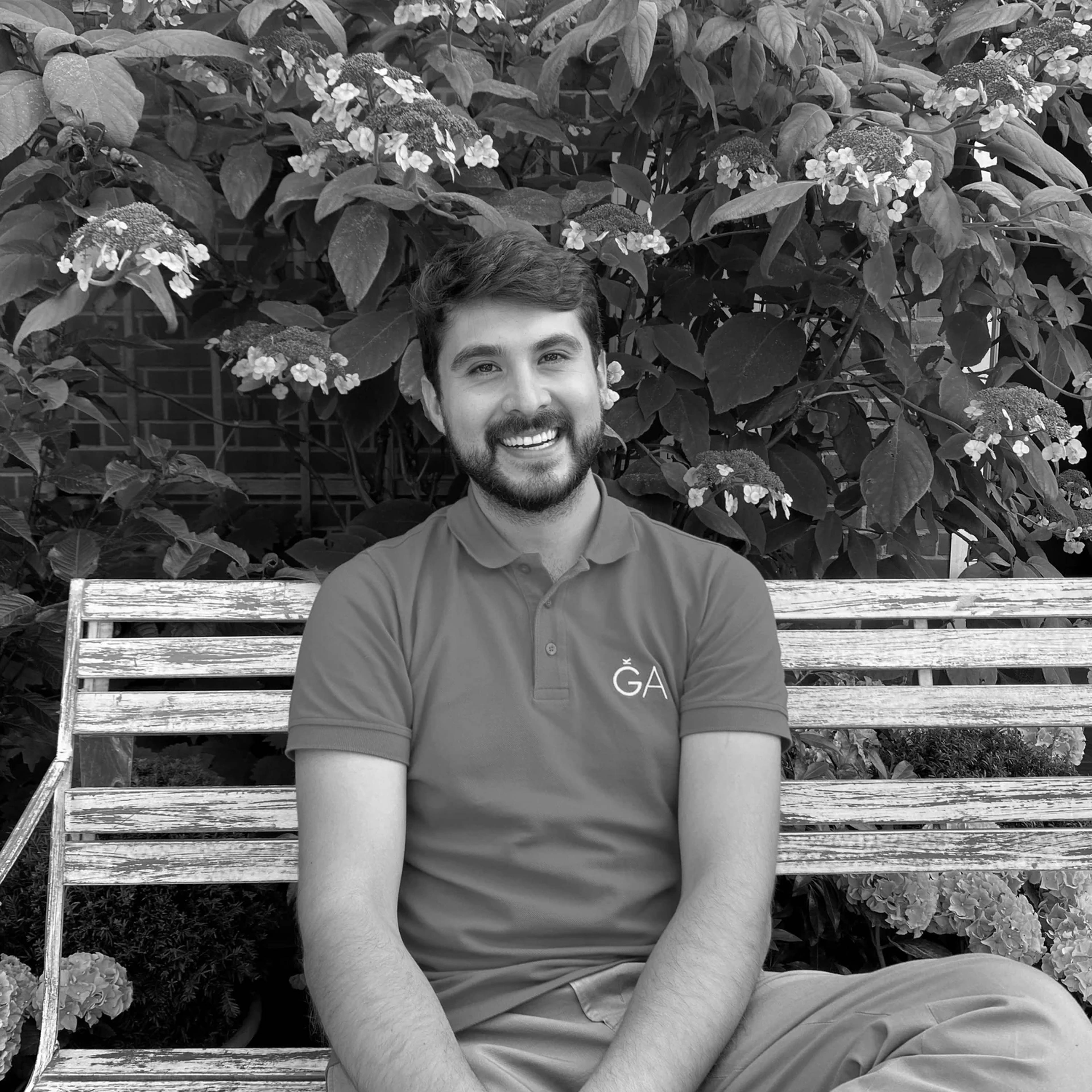 A smiling Sebastian Keller-Lewis who sits on a bench in front of a lush background of blooming hydrangeas. He is wearing a polo shirt with a ǦA logo. The photo is in black and white.