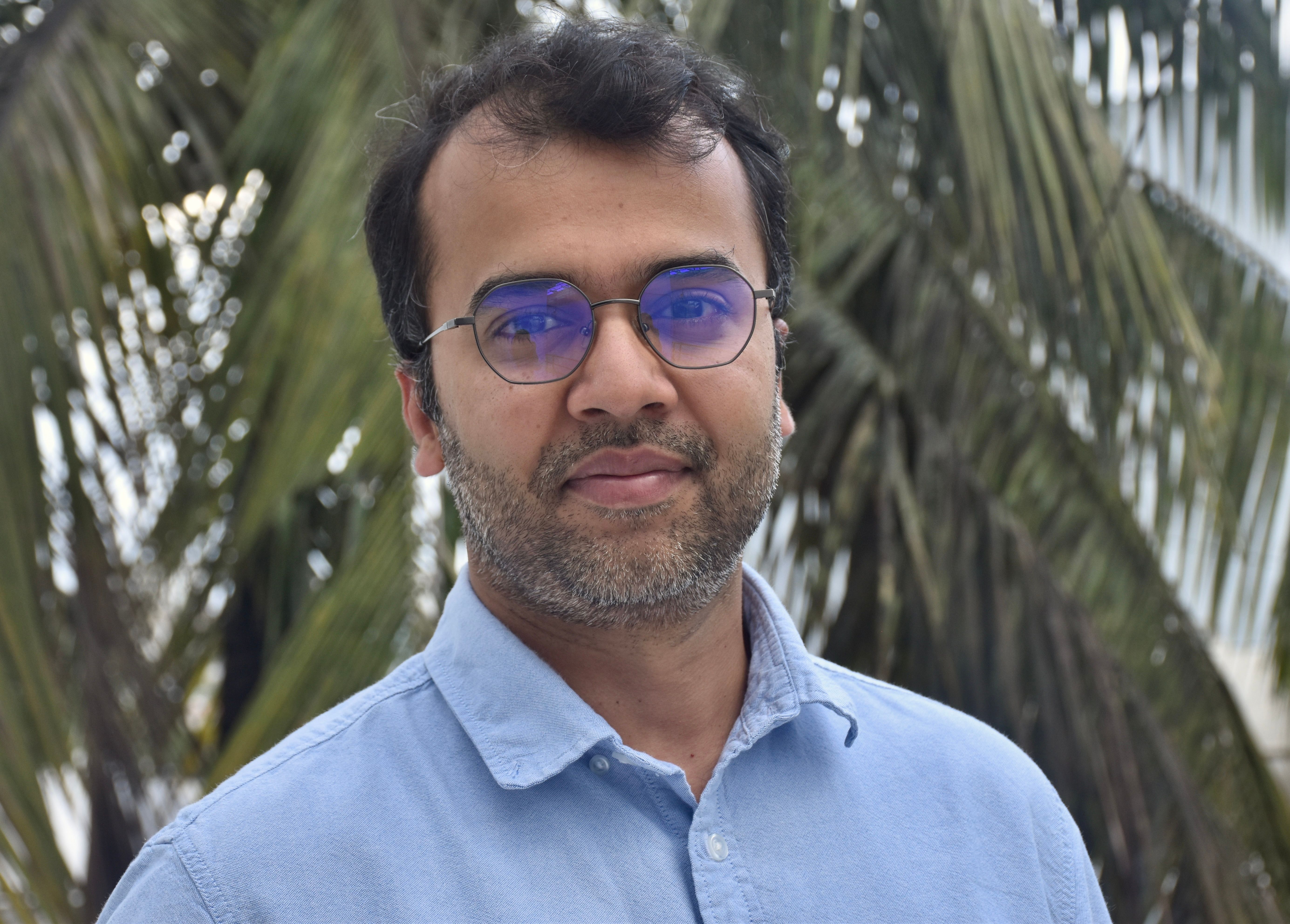 A person with short dark hair and glasses wearing a light blue shirt stands outdoors in front of green palm leaves, looking at the camera with a calm expression.