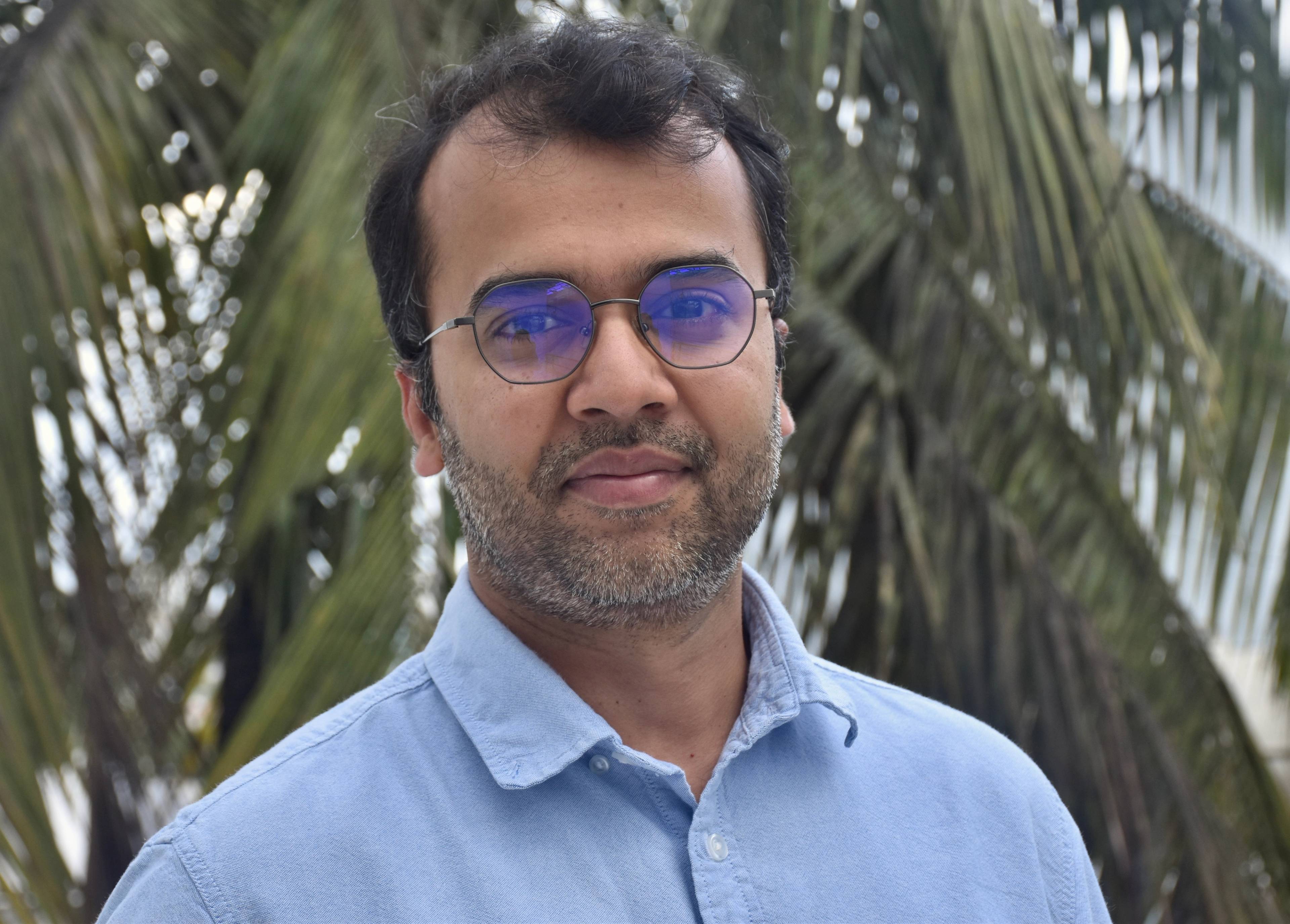 A person with short dark hair and glasses wearing a light blue shirt stands outdoors in front of green palm leaves, looking at the camera with a calm expression.