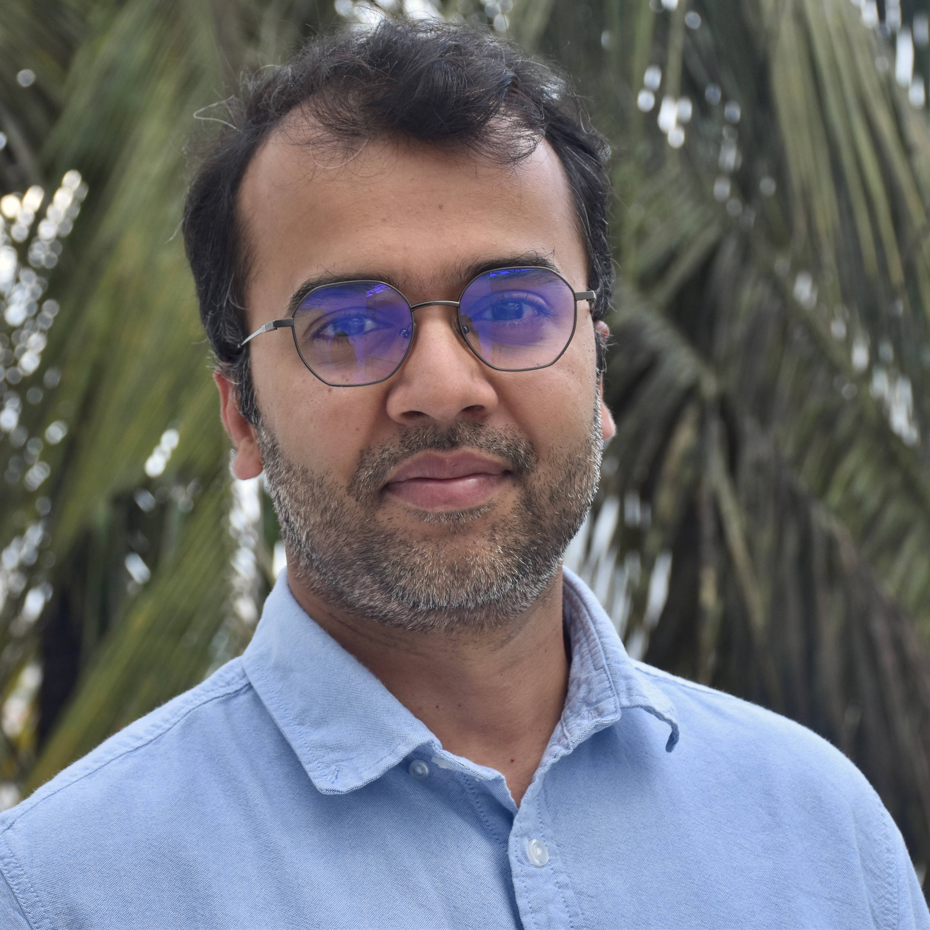 A person with short dark hair and glasses wearing a light blue shirt stands outdoors in front of green palm leaves, looking at the camera with a calm expression.