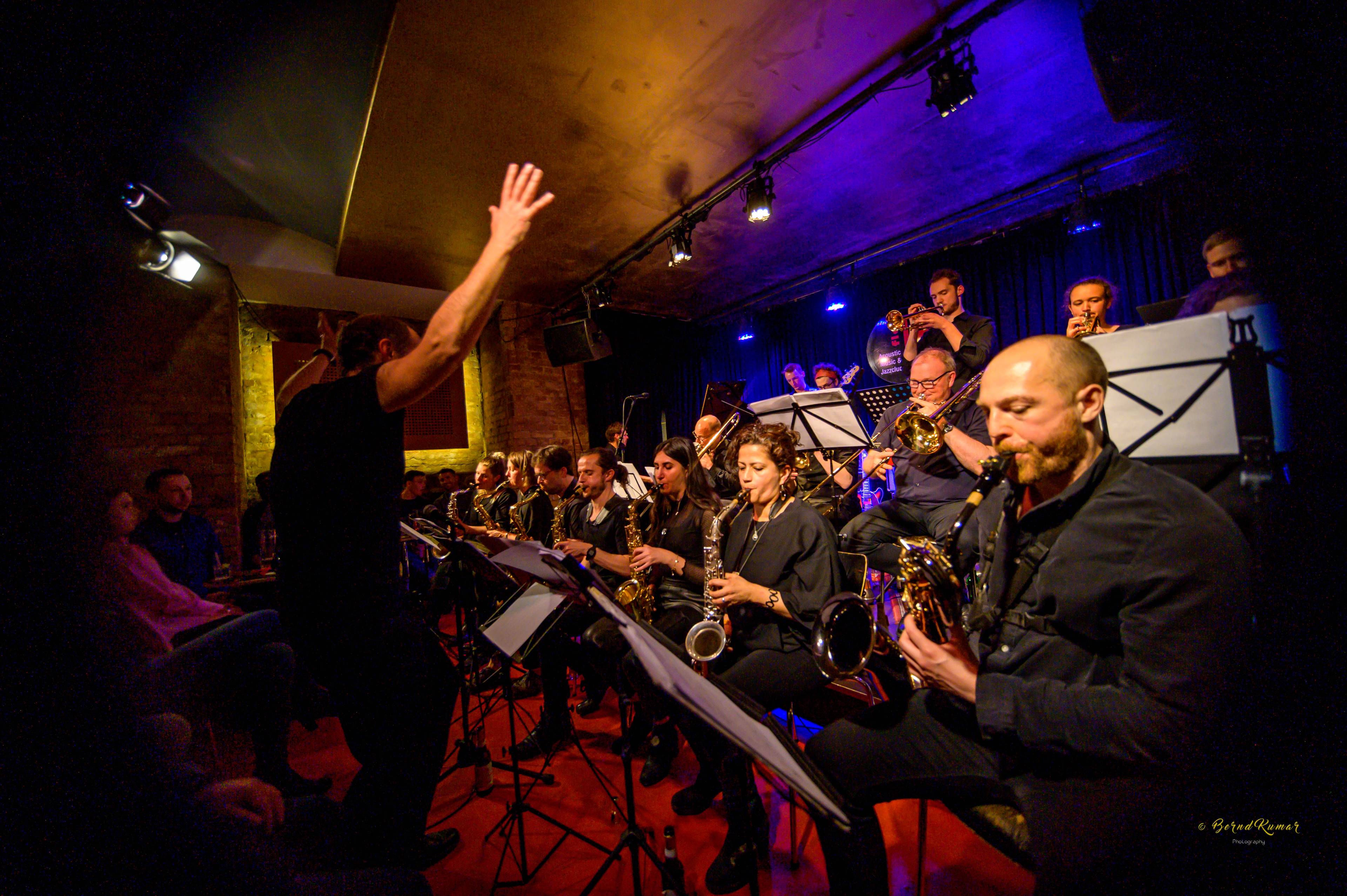 A big band performs live in a club with red flooring and a dark stage. Musicians are seated closely, playing saxophones, trumpets, and trombones, while a conductor stands in front with raised arms. The audience sits close to the performance.