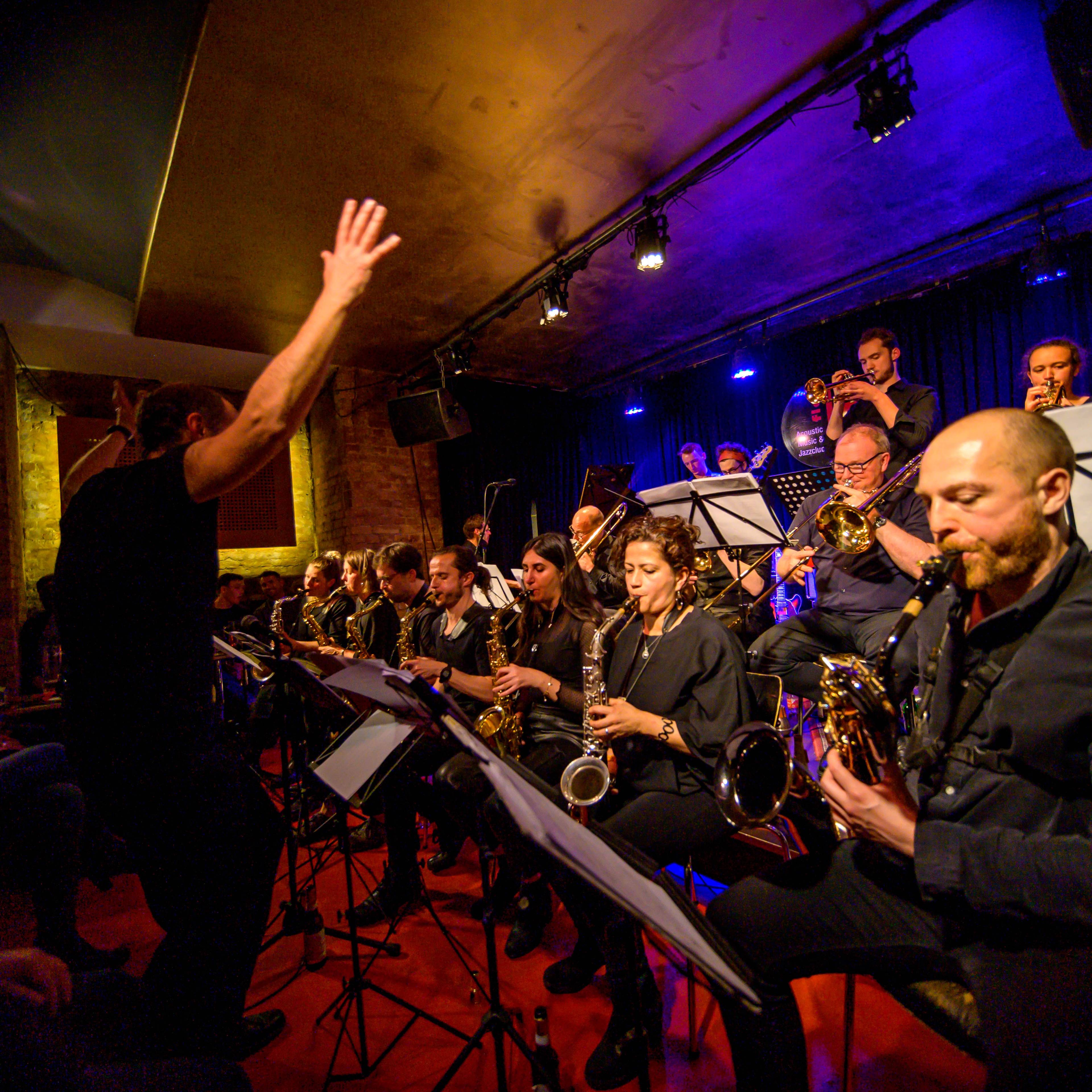 Eine Bigband spielt live in einem Club mit rotem Teppich und dunkler Bühne. Die Musiker:innen sitzen eng beisammen und spielen Saxofon, Trompete und Posaune, während eine dirigierende Person mit erhobenen Armen vorne steht. Das Publikum sitzt nah am Geschehen.