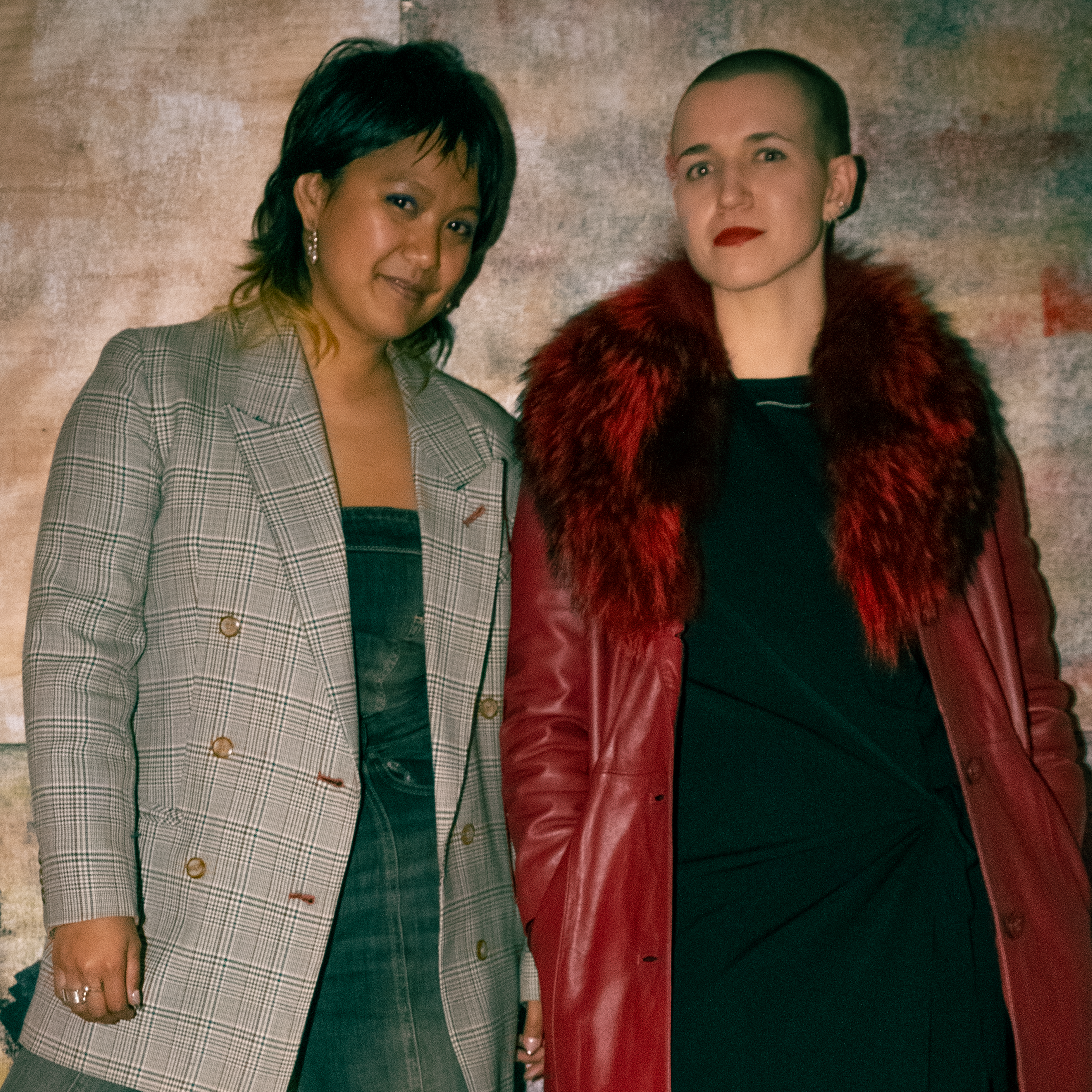 Two stylish individuals stand together. One wears a plaid coat over a dark dress, and the other wears a bold red coat with a fur collar over a black outfit. The background is an abstract, textured painting.
