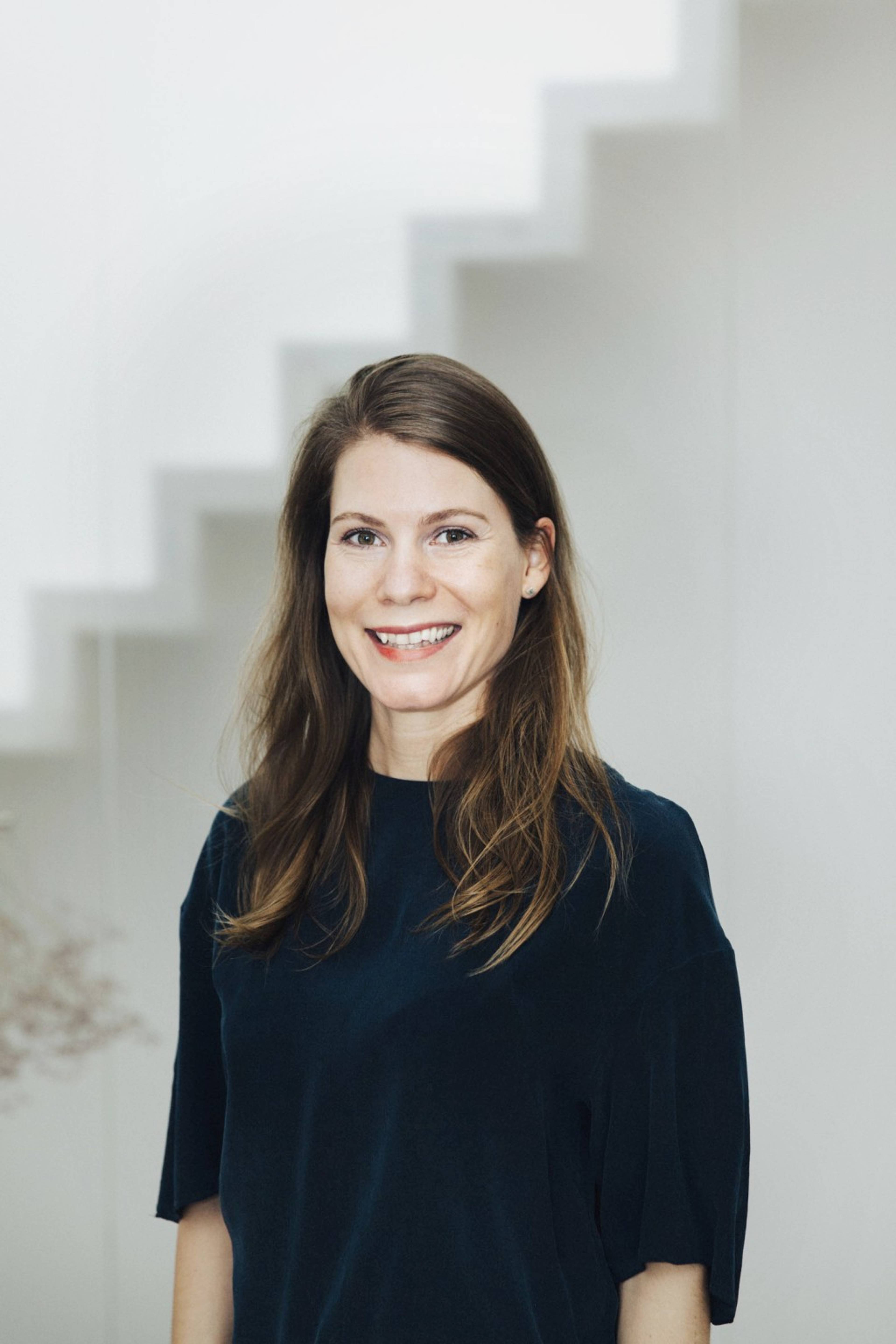 A person with long brown hair is smiling, standing indoors in front of a white staircase.