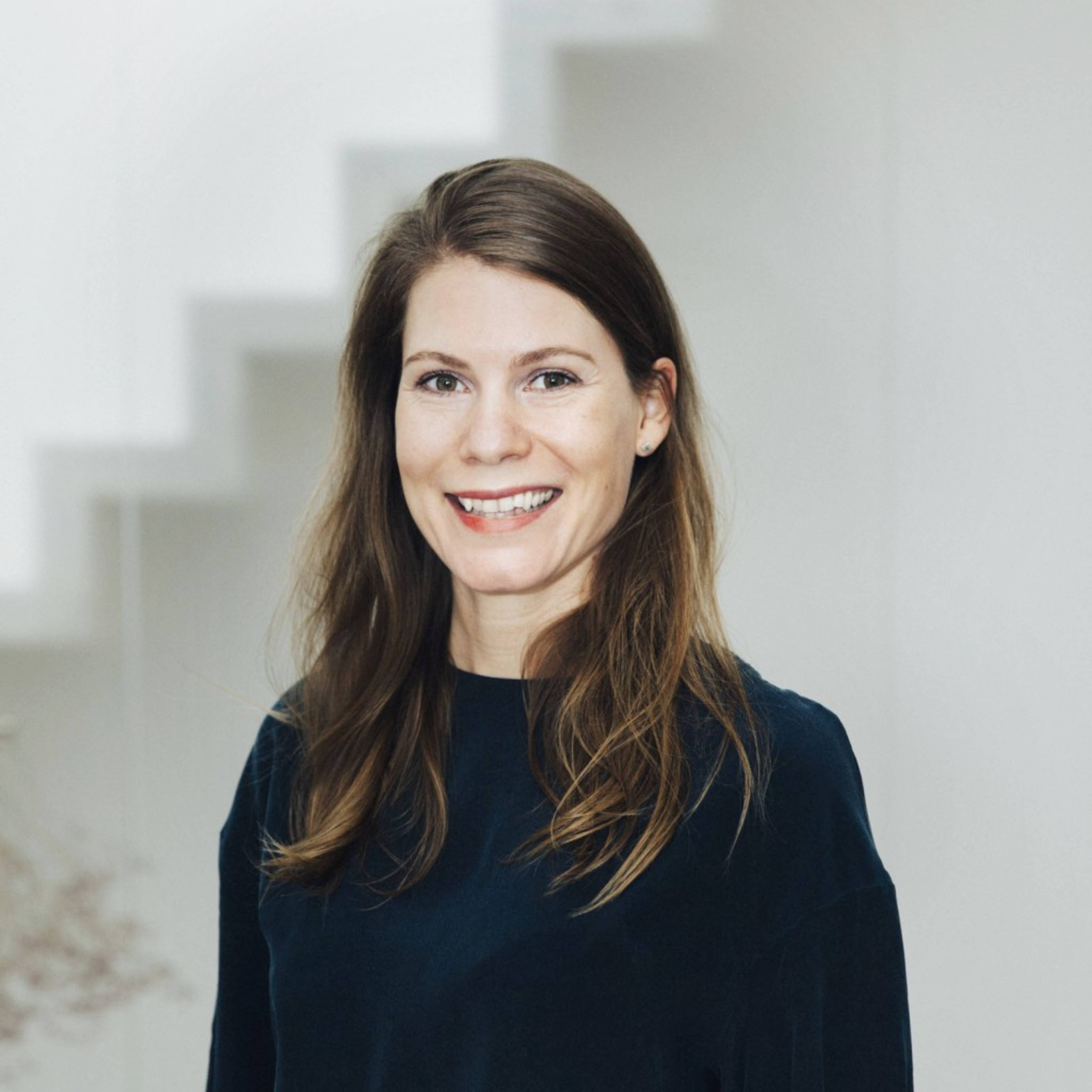 A person with long brown hair is smiling, standing indoors in front of a white staircase.