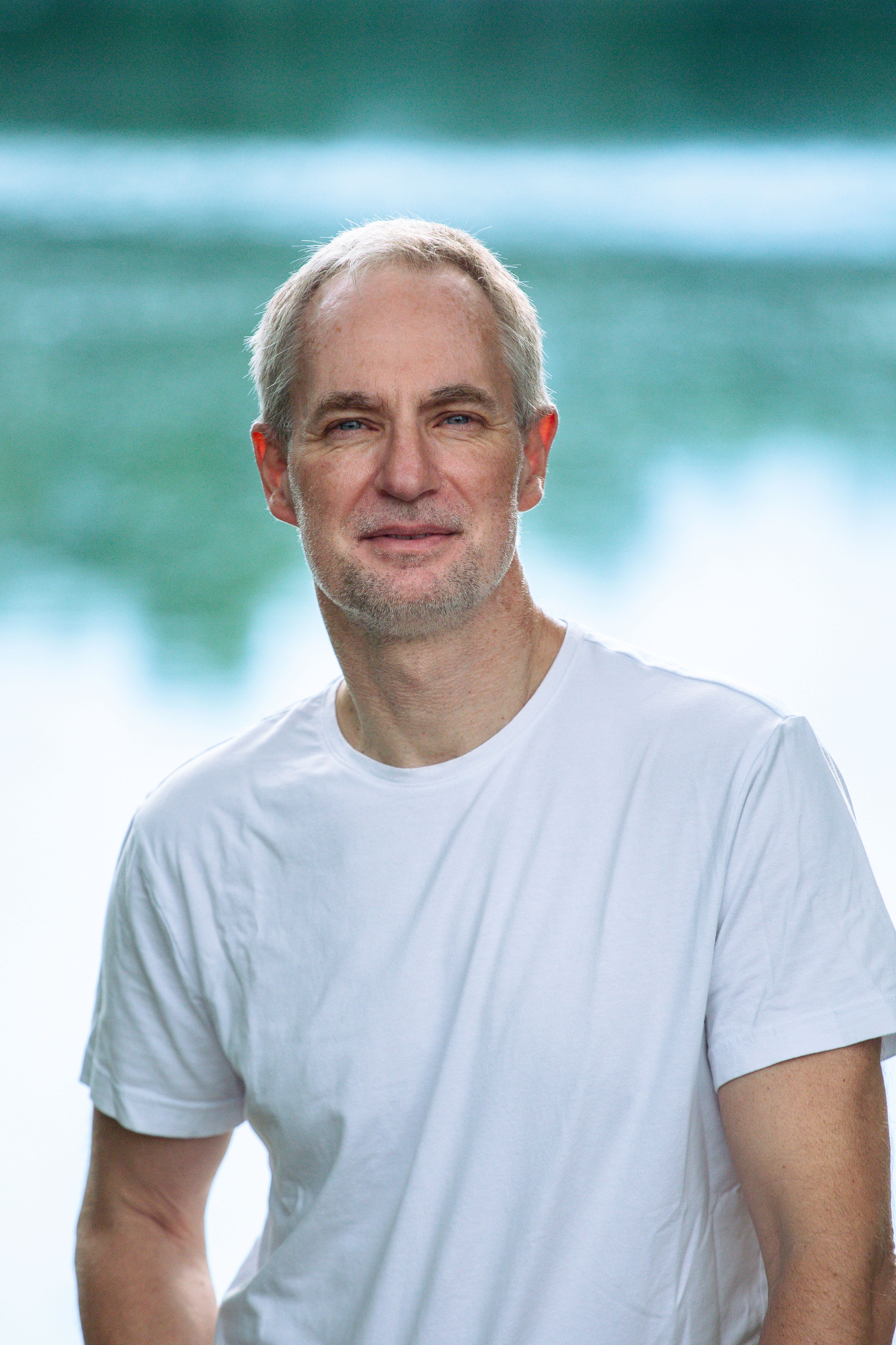 A person with short light hair wearing a plain white T-shirt stands outdoors in front of a softly blurred background of water and greenery, looking at the camera with a calm expression.