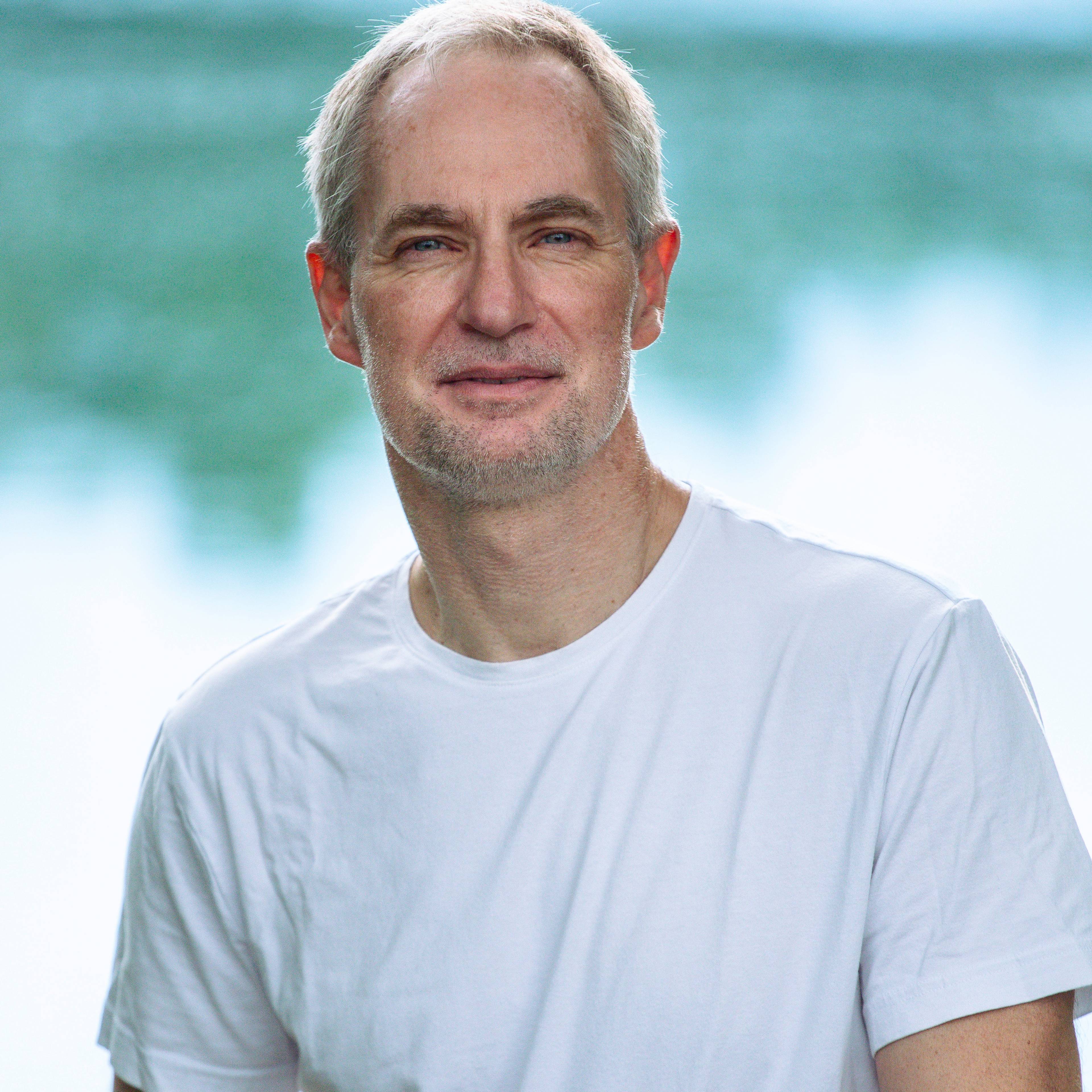 A person with short light hair wearing a plain white T-shirt stands outdoors in front of a softly blurred background of water and greenery, looking at the camera with a calm expression.