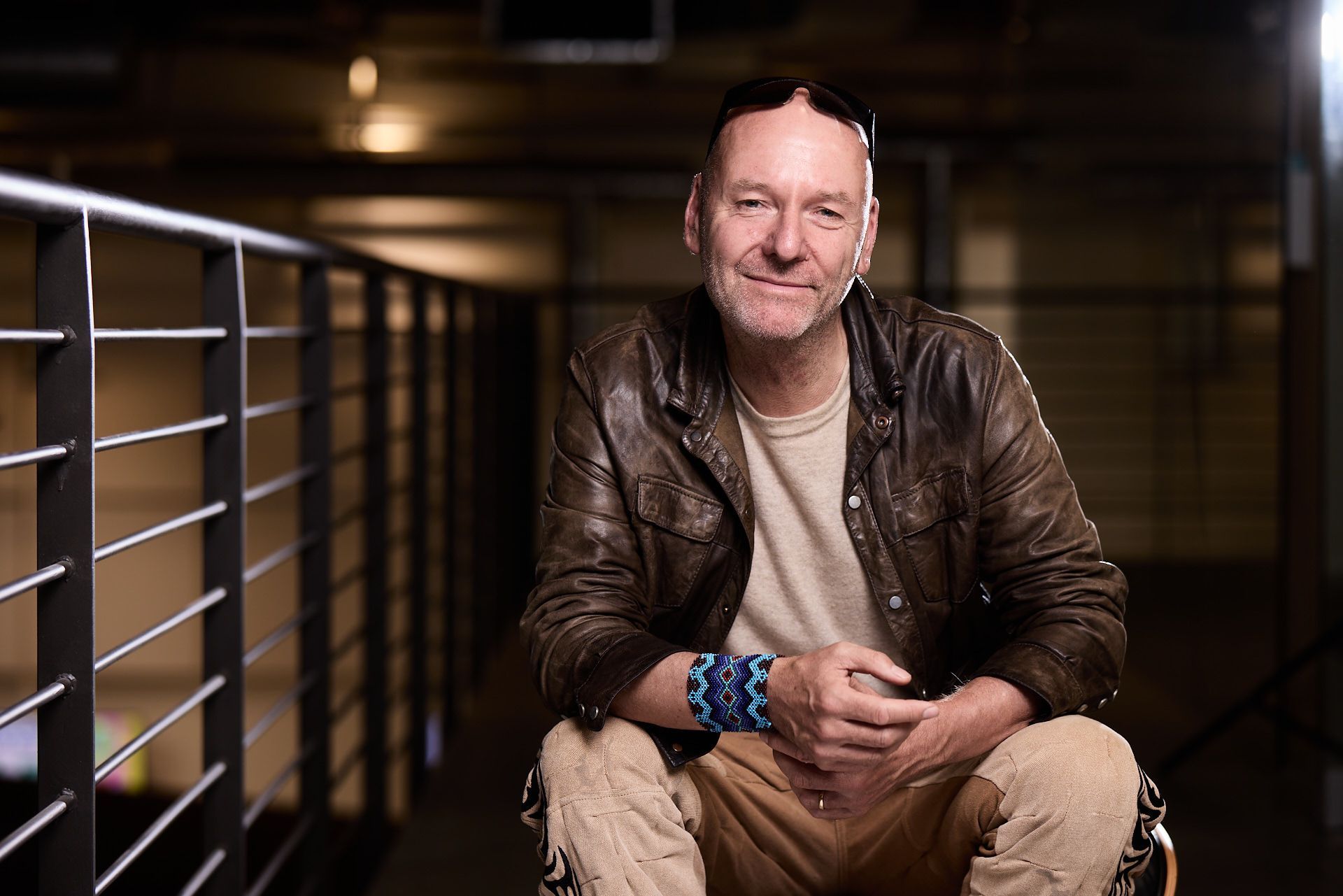 A person with short hair sits on a chair in an indoor industrial space, smiling slightly. They wear a brown leather jacket, beige pants, and a beaded blue bracelet.