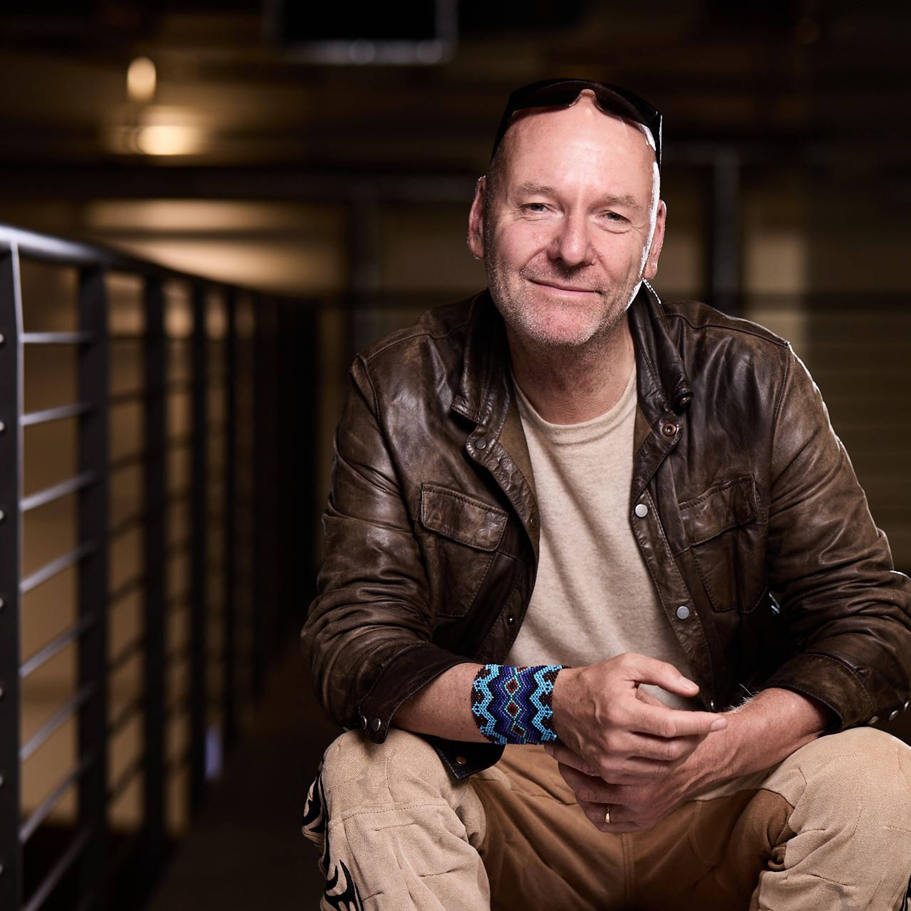 A person with short hair sits on a chair in an indoor industrial space, smiling slightly. They wear a brown leather jacket, beige pants, and a beaded blue bracelet.