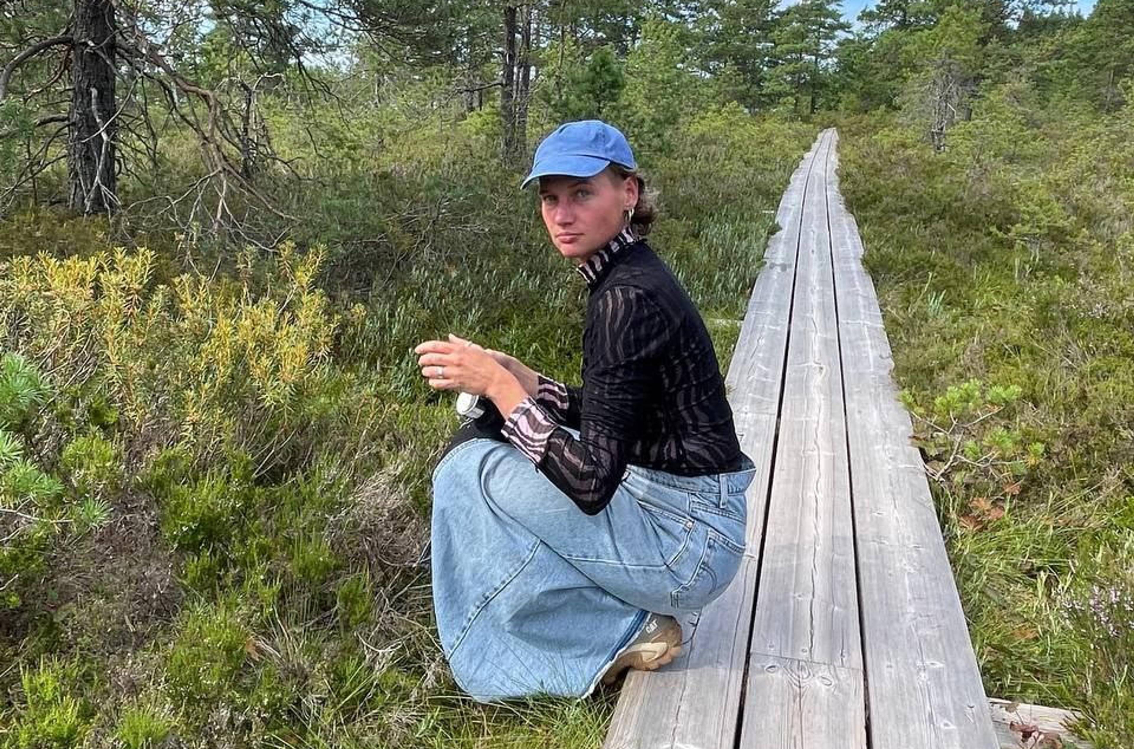 A person wearing a blue cap, black top, and wide denim skirt sits at the edge of a wooden boardwalk in the middle of a green bog landscape, looking at the camera.
