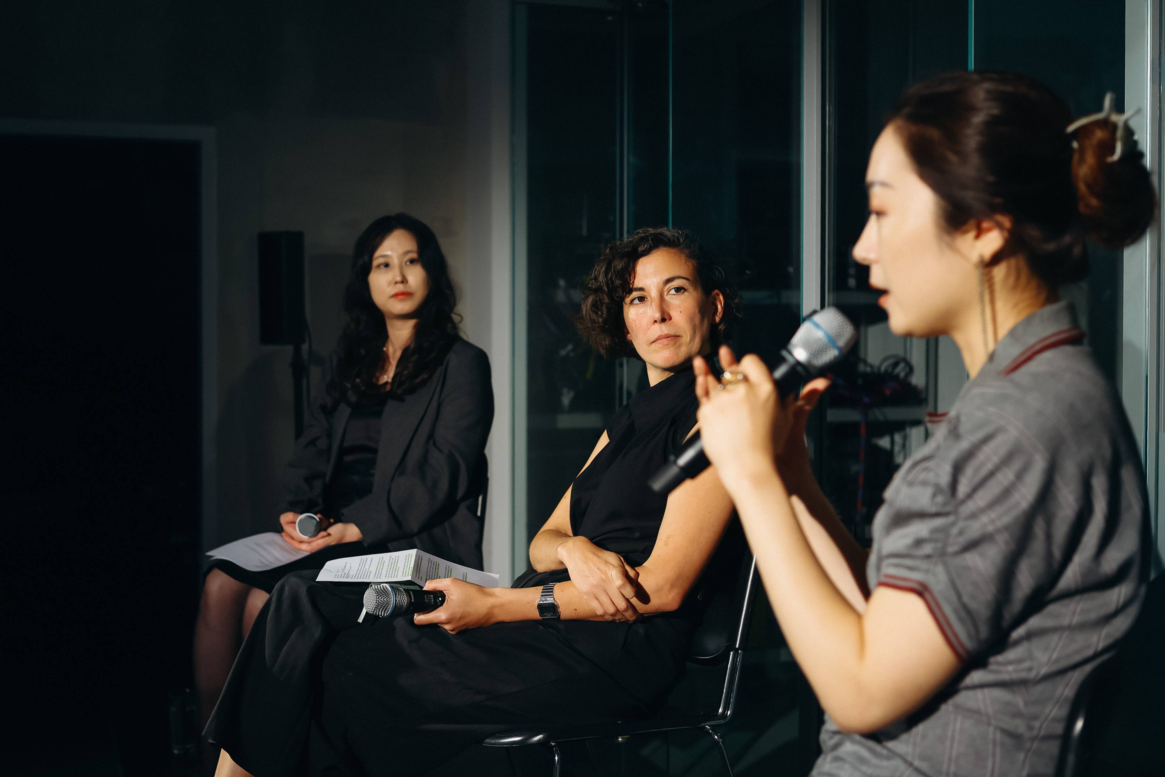 Drei Frauen sitzen während einer Podiumsdiskussion auf der Bühne. Die Frau rechts trägt ein graues Kleid, hat ihre Haare zu einem Dutt gebunden und spricht gestikulierend in ein Handmikrofon; die beiden Frauen neben ihr hören zu und halten jeweils ein Mikrofon.