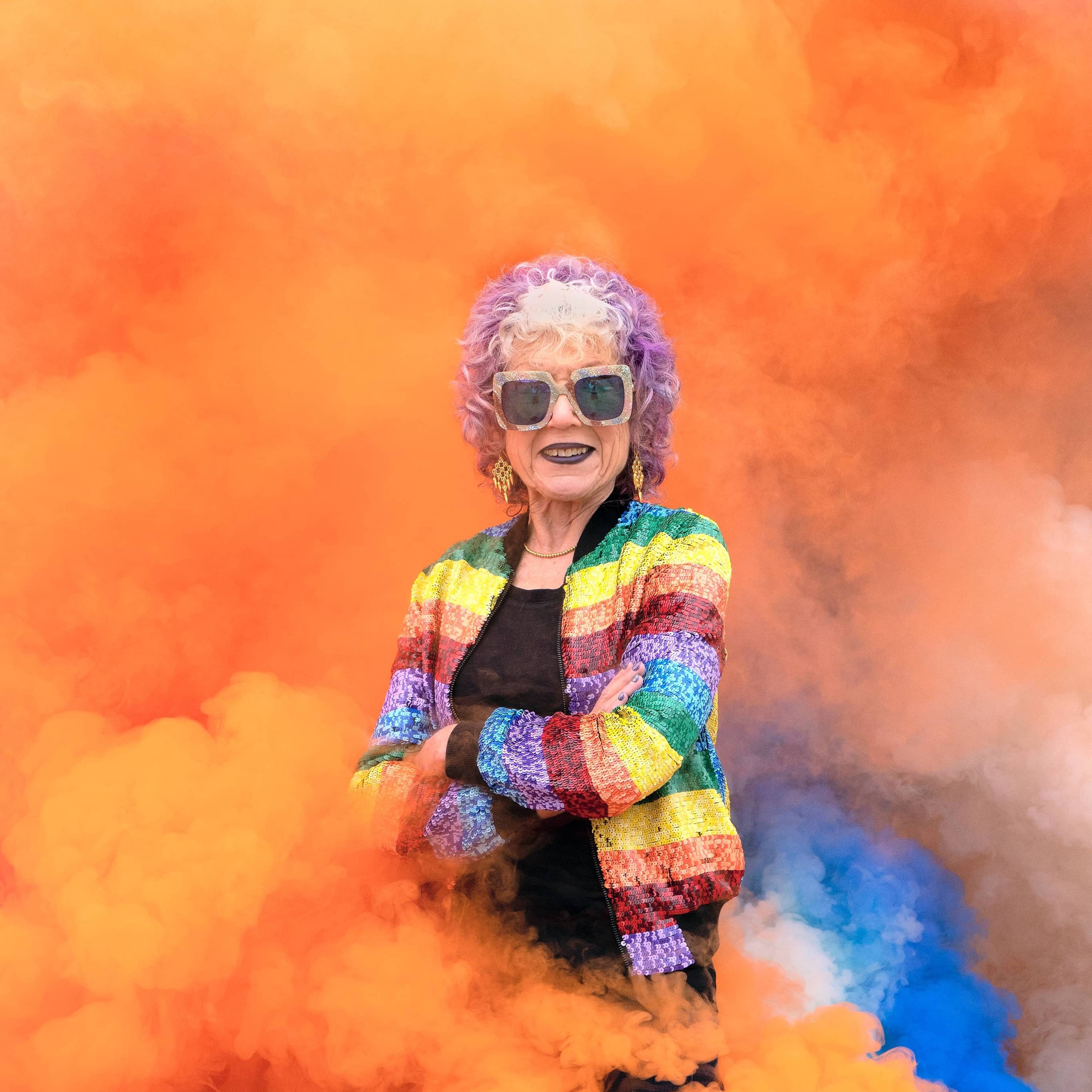 Eine ältere Frau mit lila Haaren und Sonnenbrille steht selbstbewusst inmitten von leuchtend orangefarbenem Rauch. Sie trägt eine bunte, gestreifte Jacke und lächelt mit verschränkten Armen. Der Hintergrund ist mit wirbelnden Orange- und Blautönen gefüllt.
