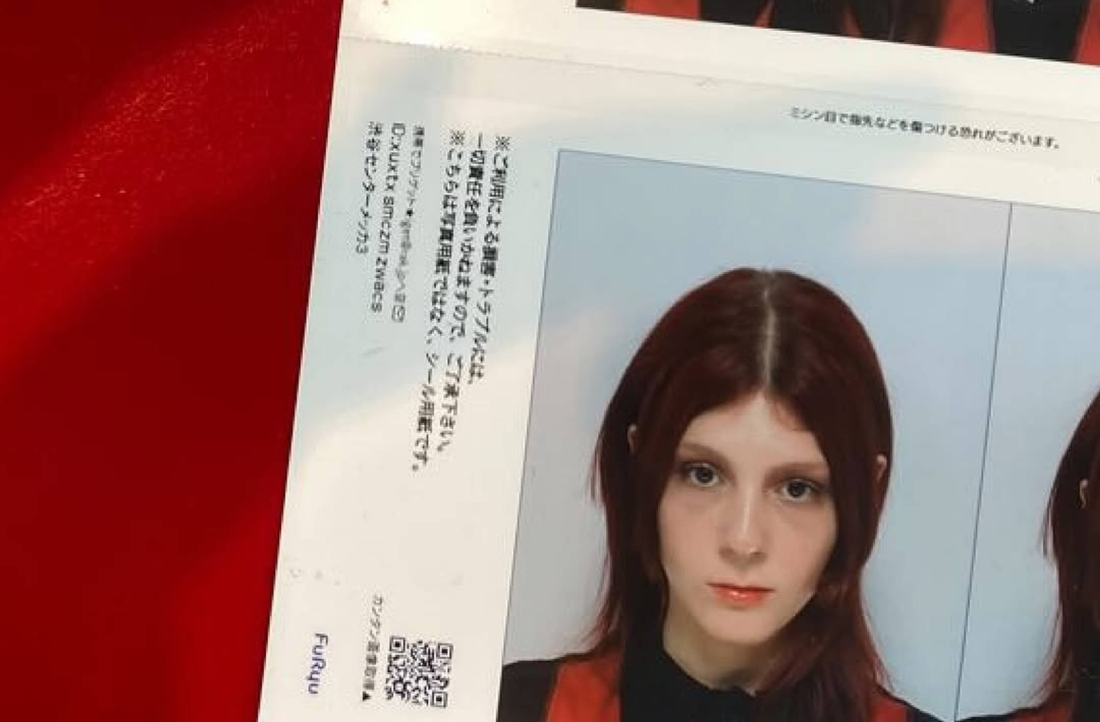 A printed photo booth sticker sheet shows a person with medium-length dark hair and a serious expression against a light background. The printout lies on a red surface, with Japanese text and a QR code visible beside the photo.