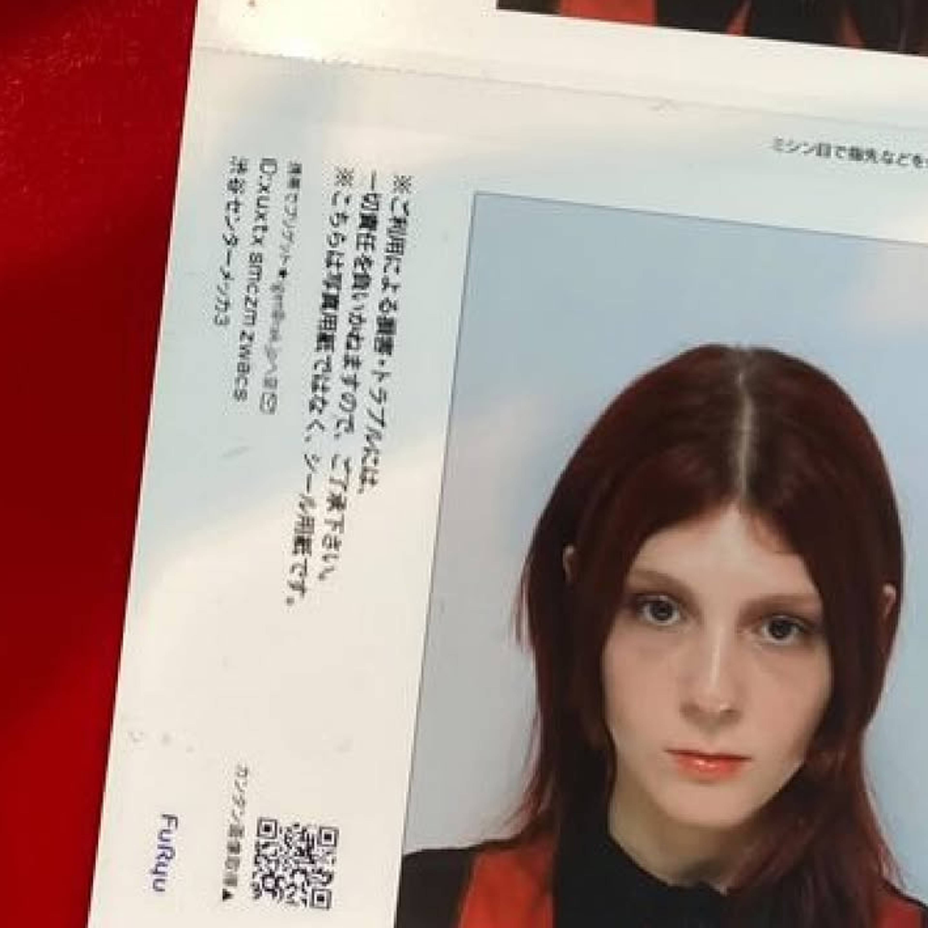A printed photo booth sticker sheet shows a person with medium-length dark hair and a serious expression against a light background. The printout lies on a red surface, with Japanese text and a QR code visible beside the photo.