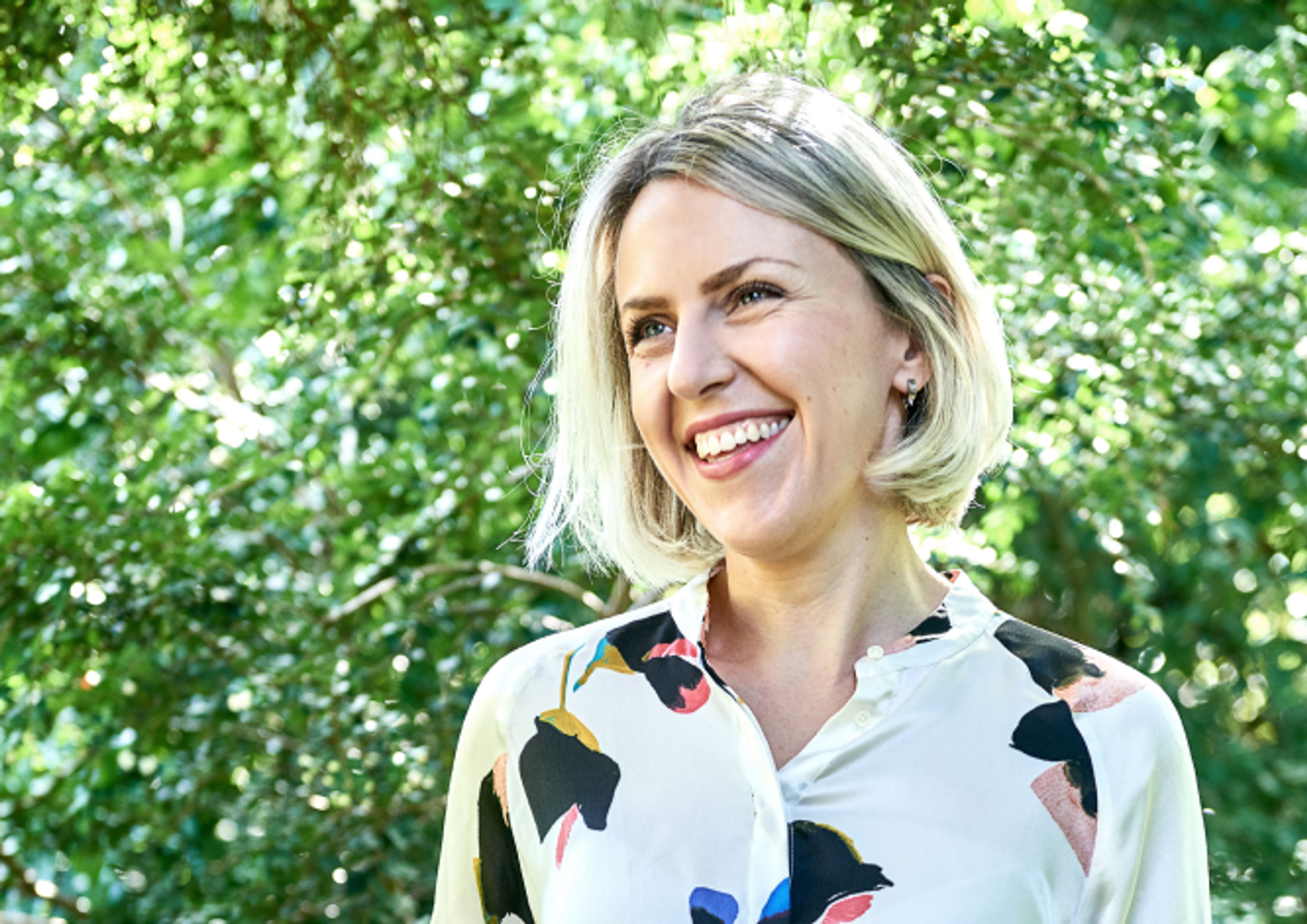 Eine lächelnde Person mit kurzen blonden Haaren steht im Freien vor üppigem grünen Laub. Sie trägt eine helle Bluse mit einem bunten Blumenmuster.