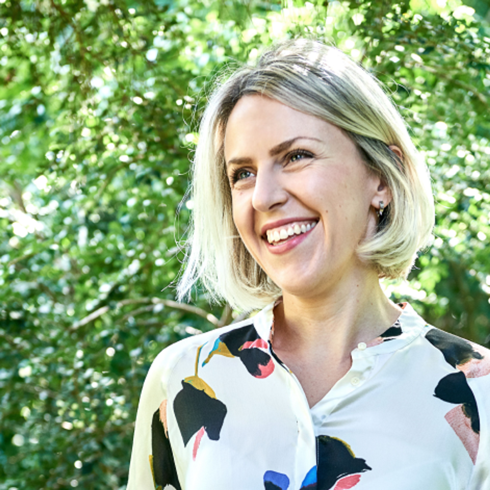 A smiling person with short blonde hair stands outdoors in front of lush green foliage. They are wearing a light-colored blouse with a colorful floral pattern.
