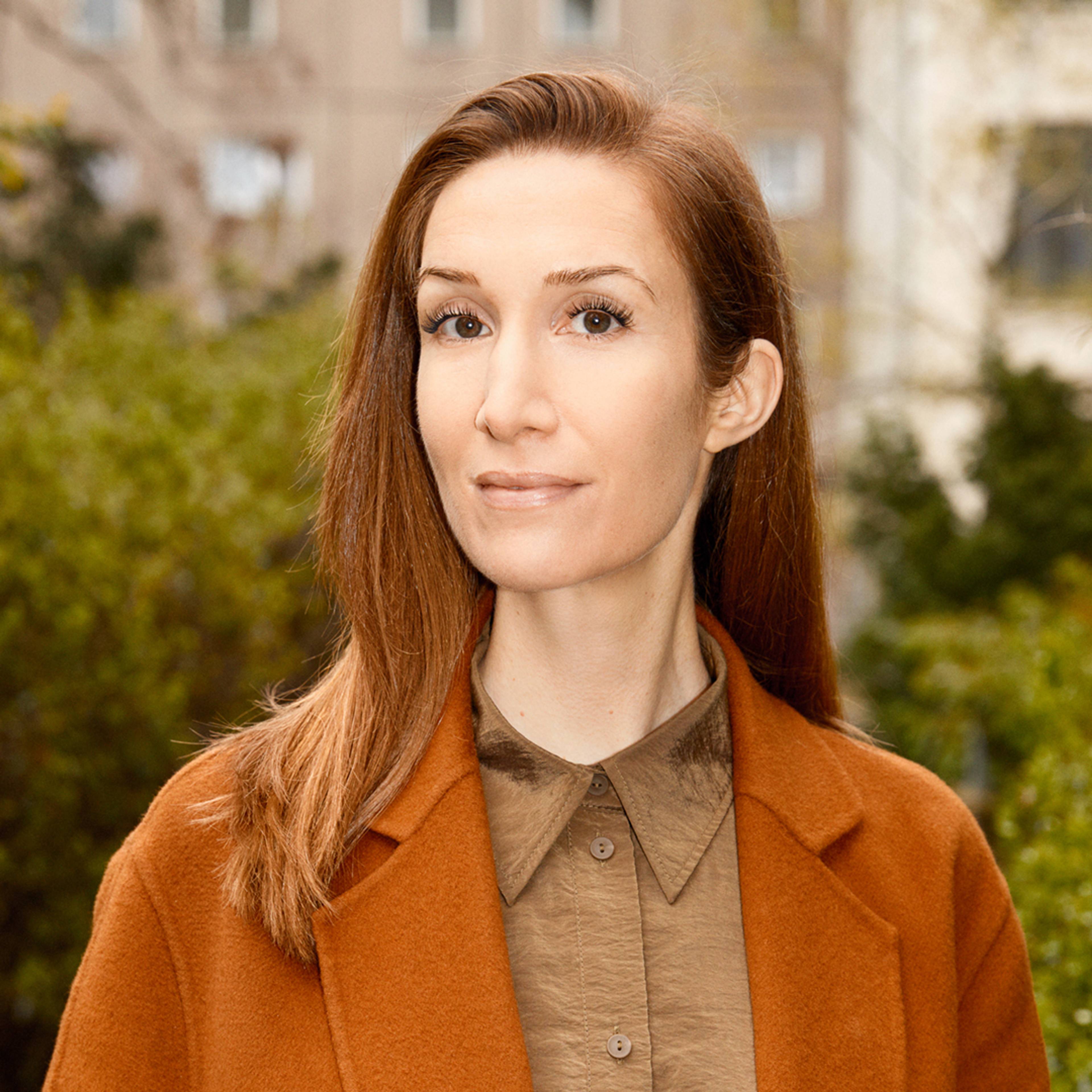 Portrait of Carly Whitefield, Head of Programme at LAS Art Foundation, standing outdoors in a rust-coloured coat and beige shirt, with greenery and urban architecture softly blurred in the background.