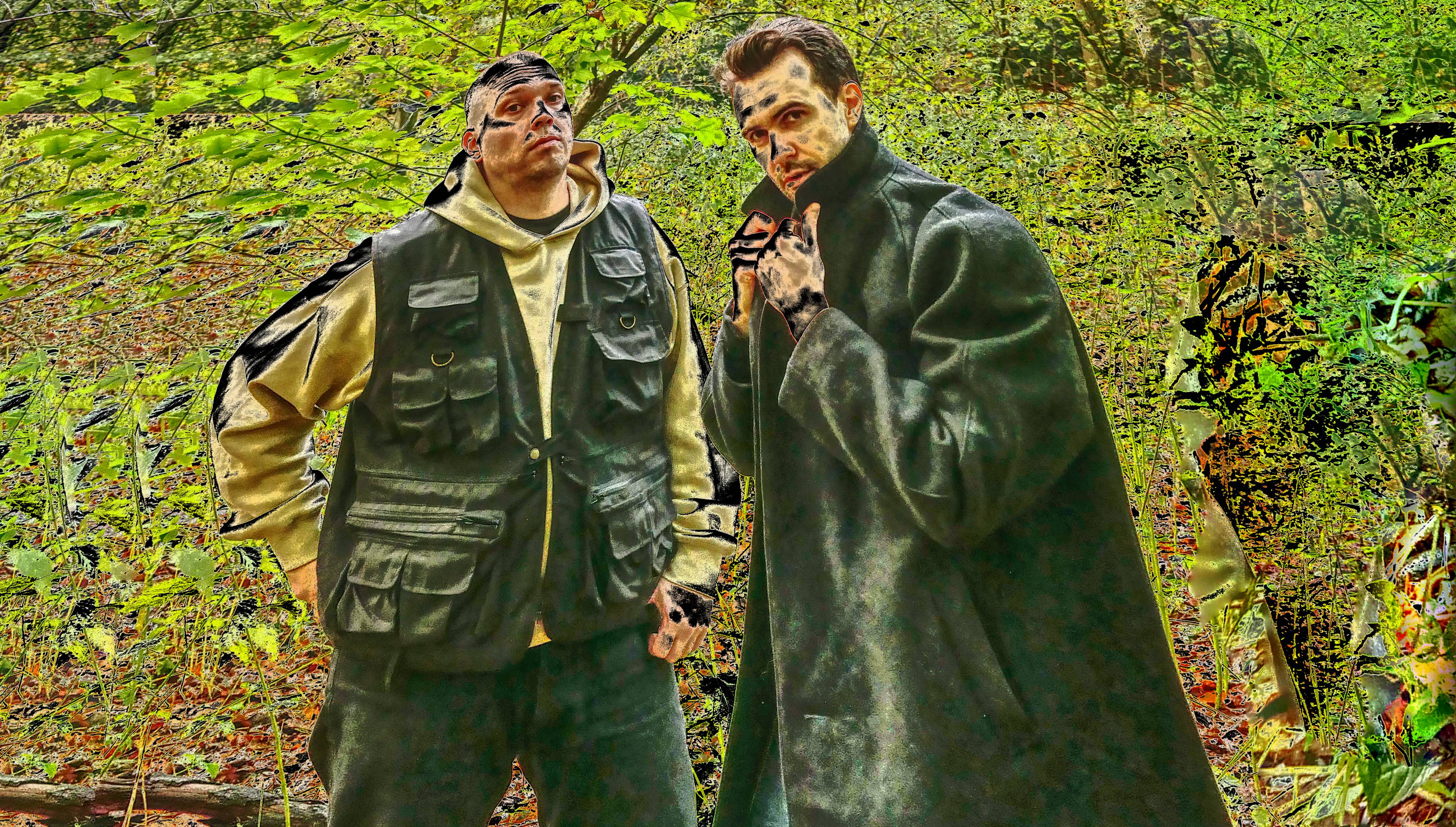 Zwei Personen stehen in einem bewaldeten Gebiet mit hellgrünem Laub. Die eine trägt eine dunkle Weste über einem Kapuzenpulli und einer Mütze, die andere einen weiten Mantel mit hochgezogenem Kragen und nimmt eine dramatische Pose ein.