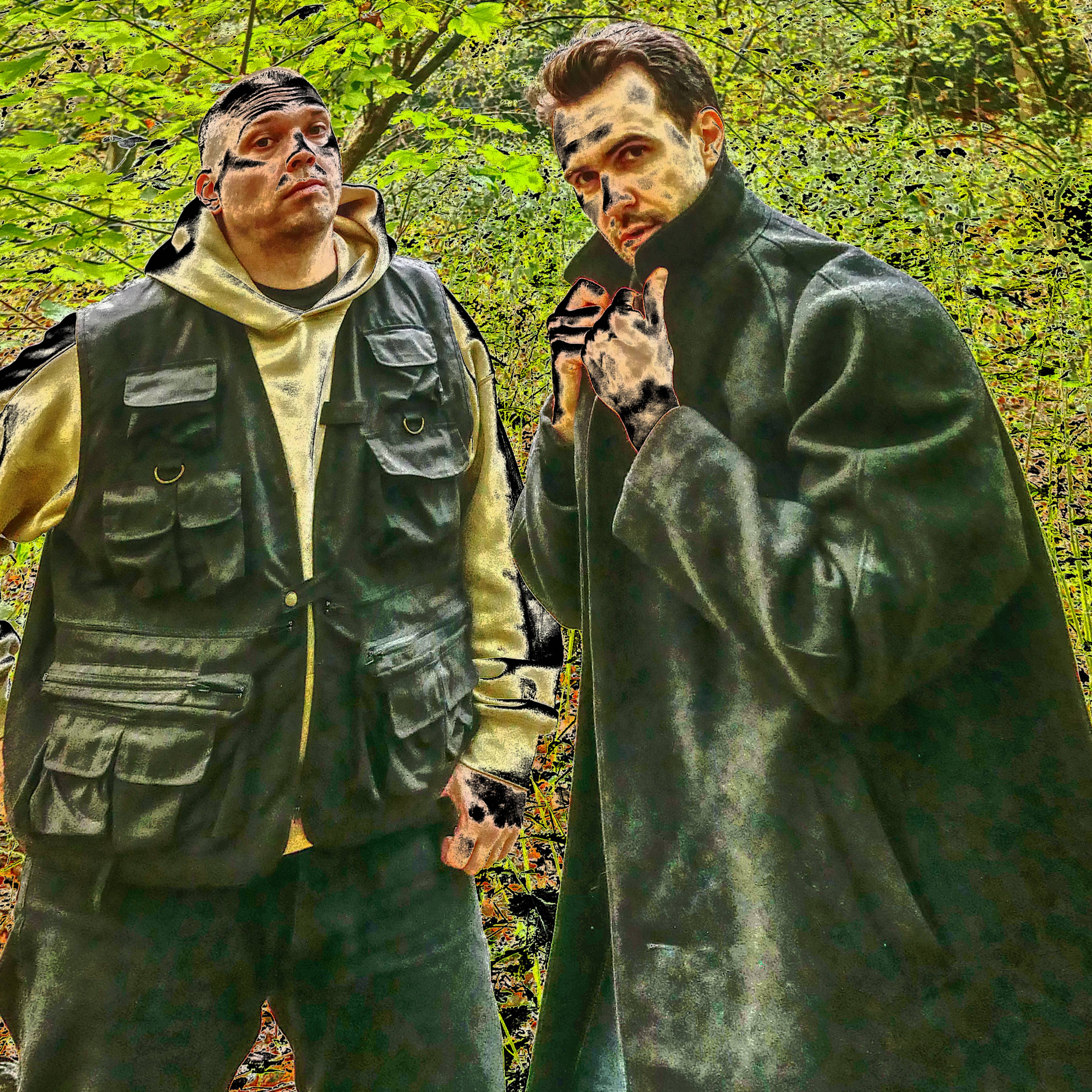 Zwei Personen stehen in einem bewaldeten Gebiet mit hellgrünem Laub. Die eine trägt eine dunkle Weste über einem Kapuzenpulli und einer Mütze, die andere einen weiten Mantel mit hochgezogenem Kragen und nimmt eine dramatische Pose ein.