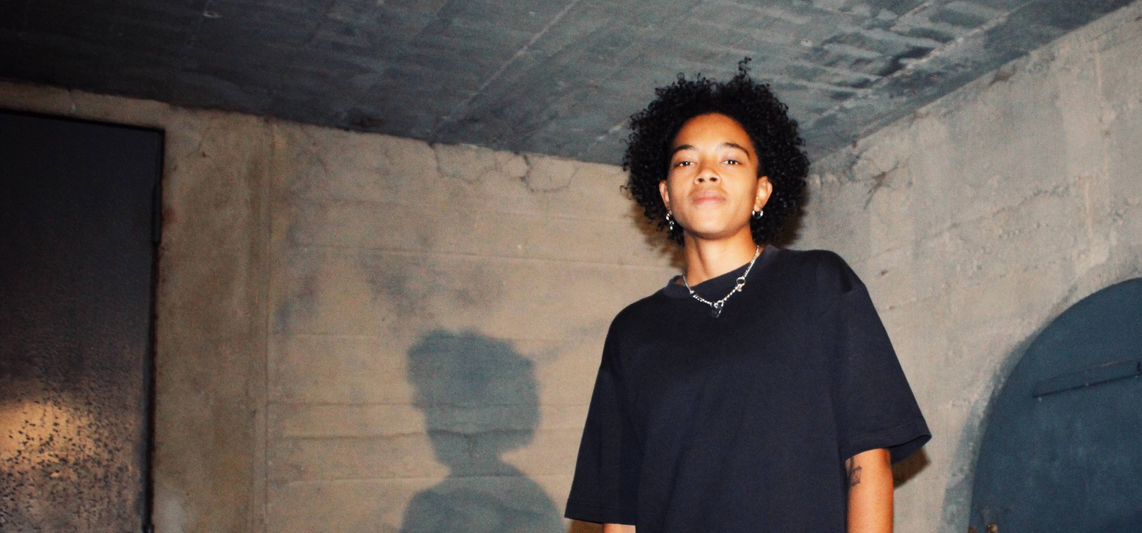 A person with curly hair and wearing a black shirt stands against a textured concrete wall. The person is looking at the camera, and their shadow is visible on the wall. The setting has an industrial, unfinished appearance.