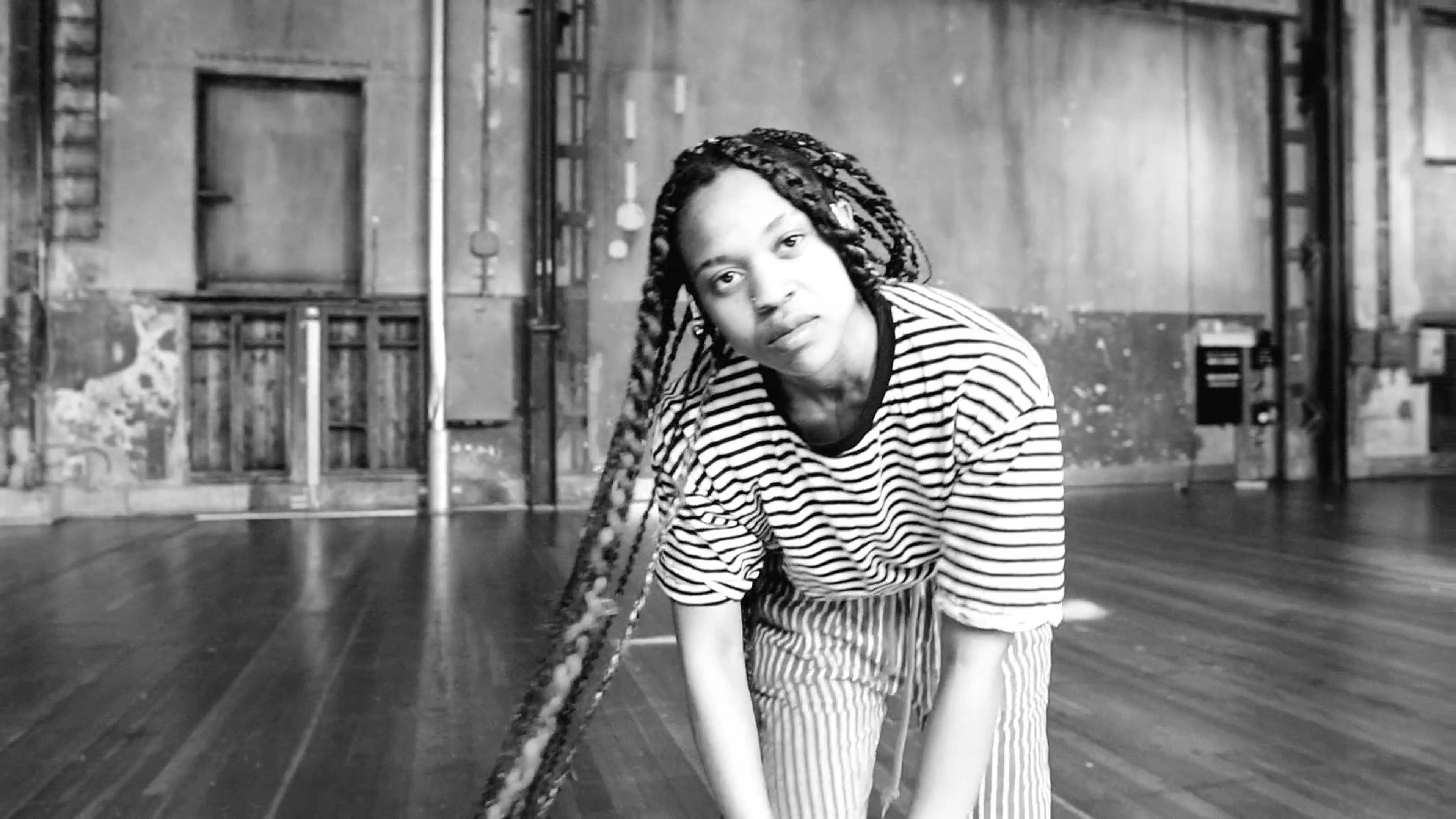 A person with long braided hair, wearing a striped shirt and pants, leans forward in an empty industrial-style room with wooden floors and weathered walls. The atmosphere is artistic and minimalist.