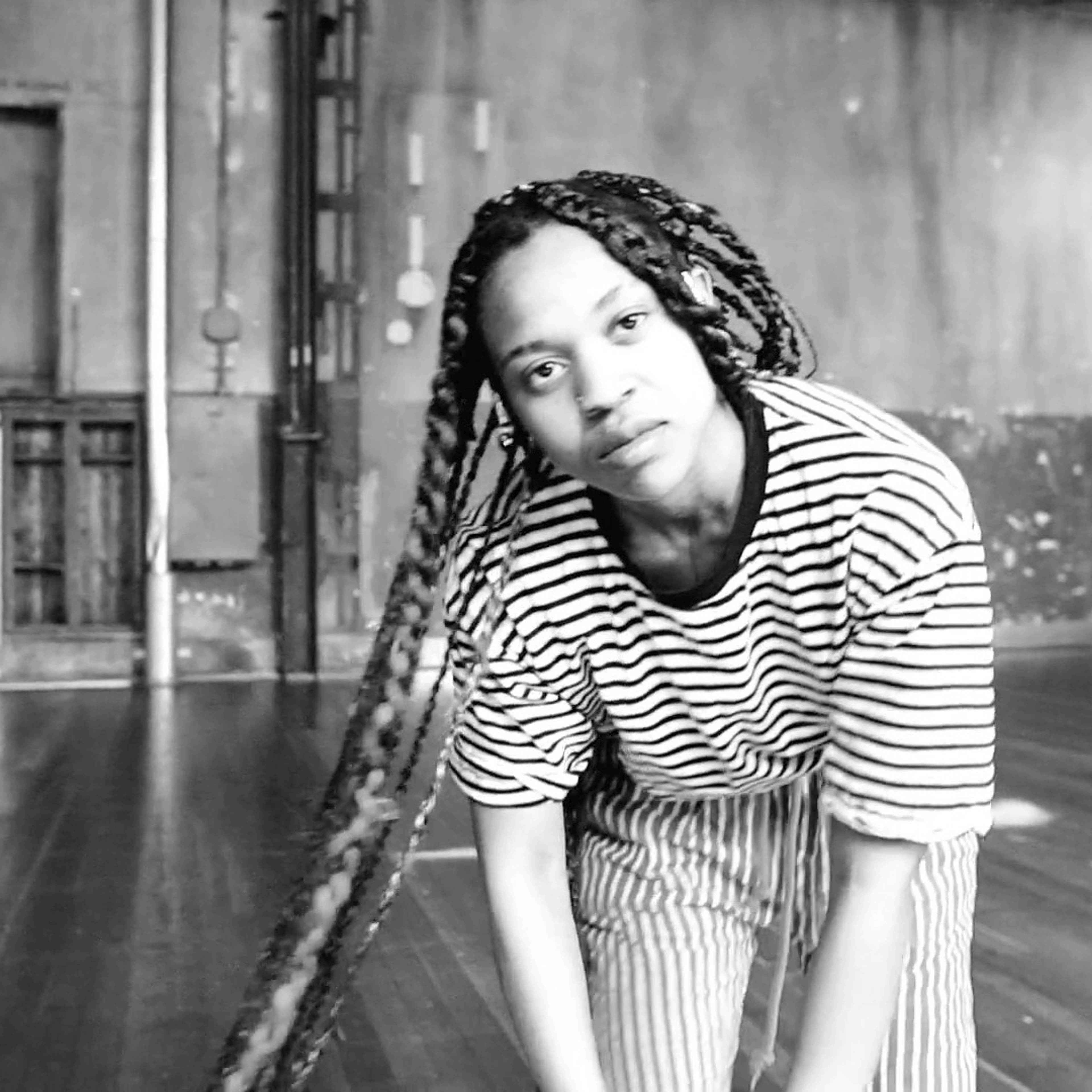 A person with long braided hair, wearing a striped shirt and pants, leans forward in an empty industrial-style room with wooden floors and weathered walls. The atmosphere is artistic and minimalist.