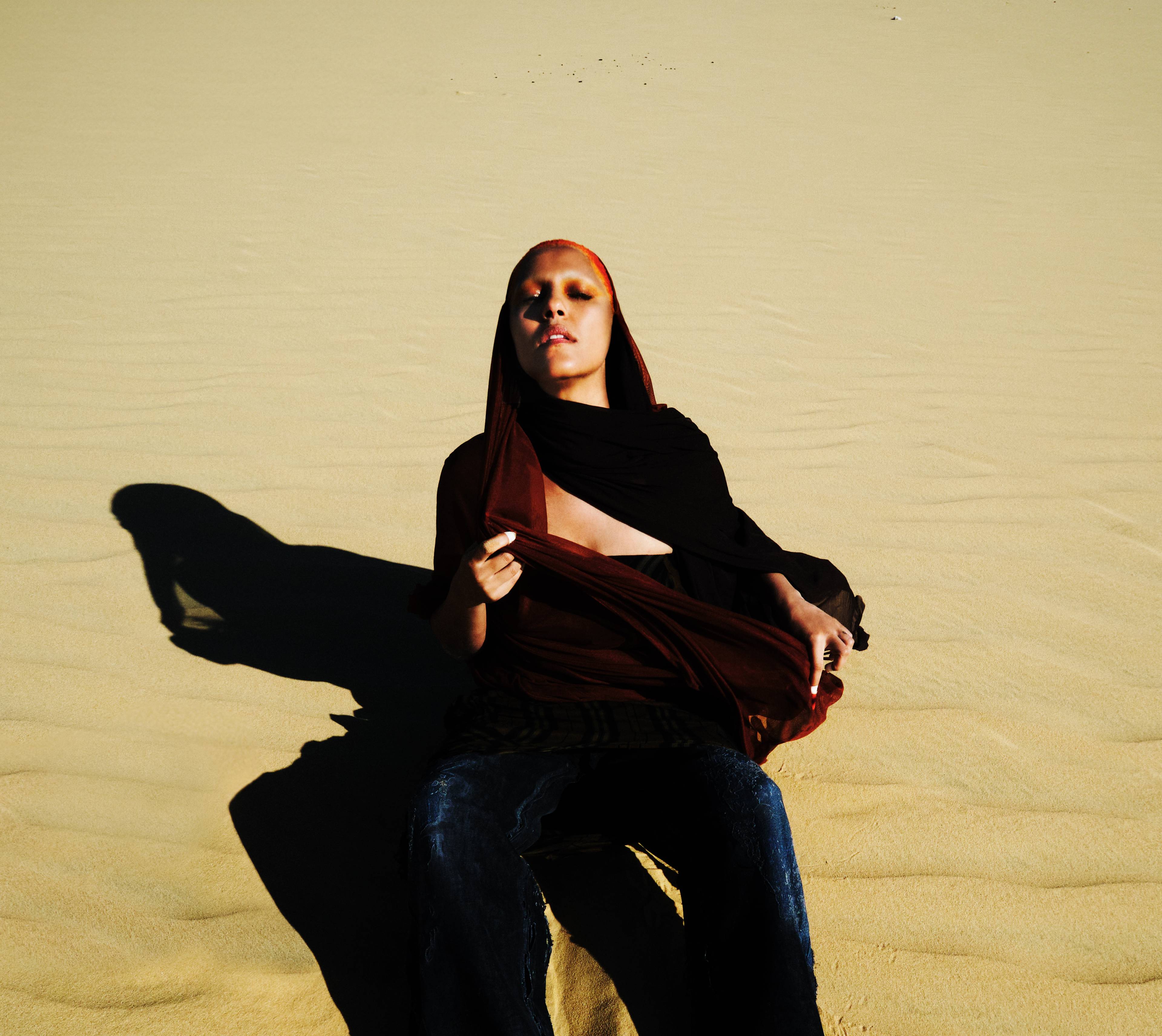 A person sits on pale desert sand with eyes closed, pulling up a cloth that partially covers their torso. Long shadows stretch across the ground, creating a powerful and contemplative atmosphere.