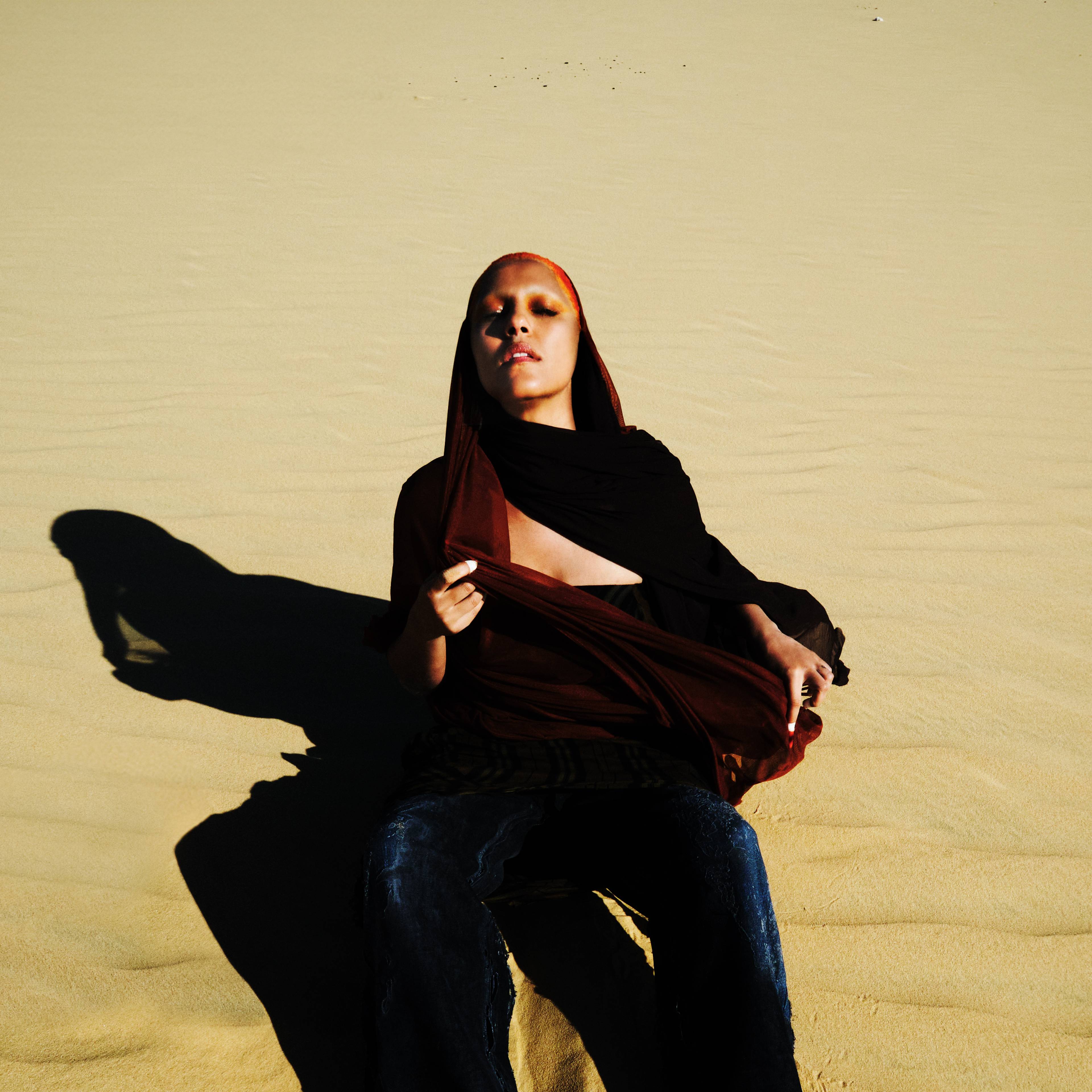 Eine Person sitzt in der Wüste auf hellem Sand, mit geschlossenen Augen und hochgezogenem Tuch, das den Oberkörper teilweise bedeckt. Lange Schatten fallen auf den Boden, die Szene wirkt kraftvoll und kontemplativ.