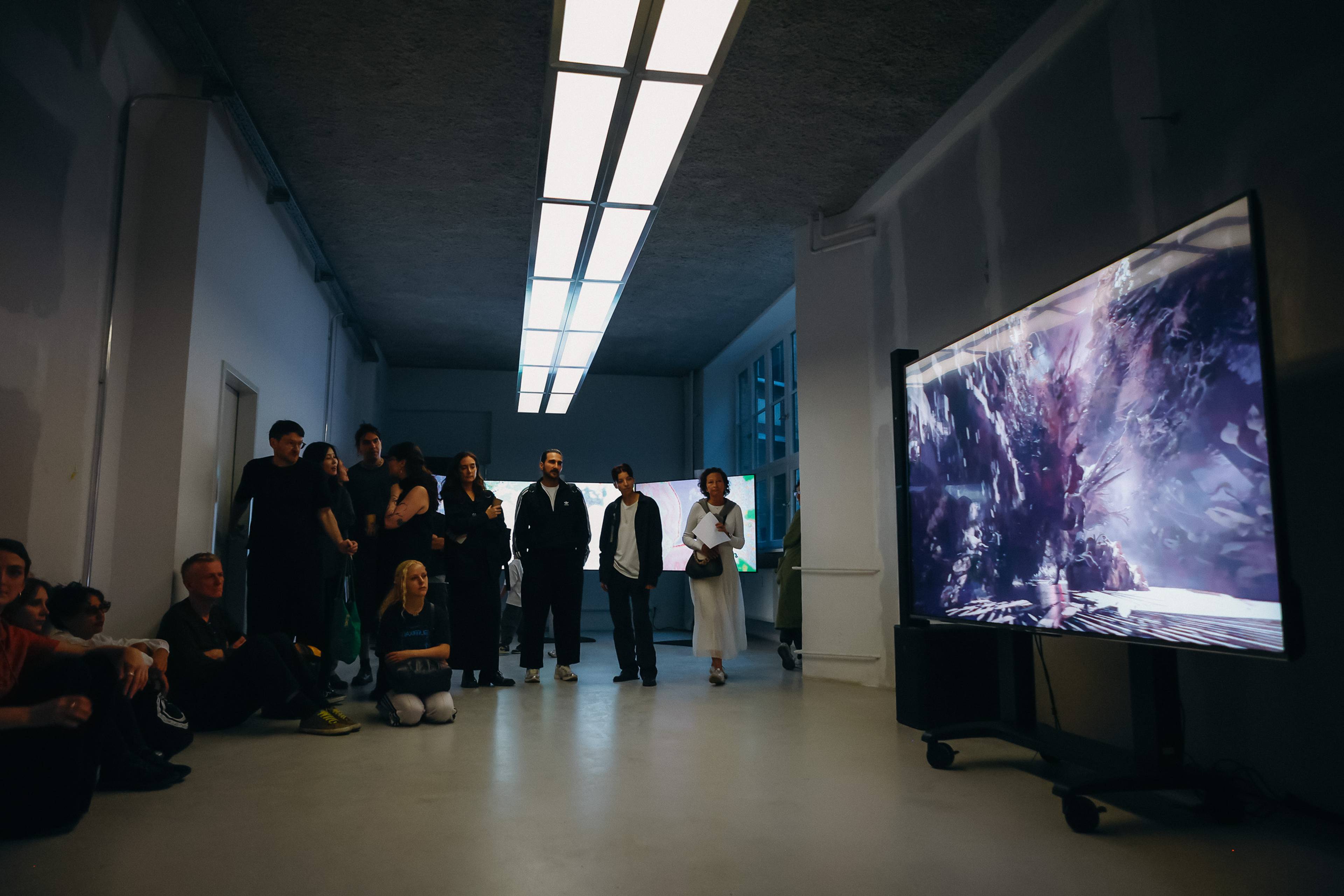 Eine Gruppe von Besucher:innen steht und sitzt in einem galerieartigen Raum und betrachtet einen großen Bildschirm, auf dem eine dunkle, abstrakte digitale Szene zu sehen ist. Im Hintergrund sind weitere Bildschirme mit farbenfrohen digitalen Bildern sichtbar.